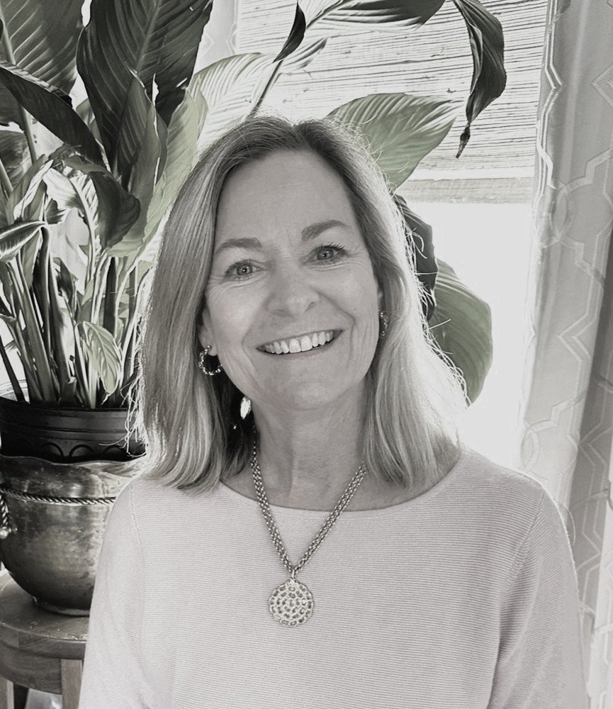 Smiling woman with shoulder-length hair wearing a light-colored top, large earrings, and a decorative pendant necklace, standing near a large leafy houseplant and a window with curtains.