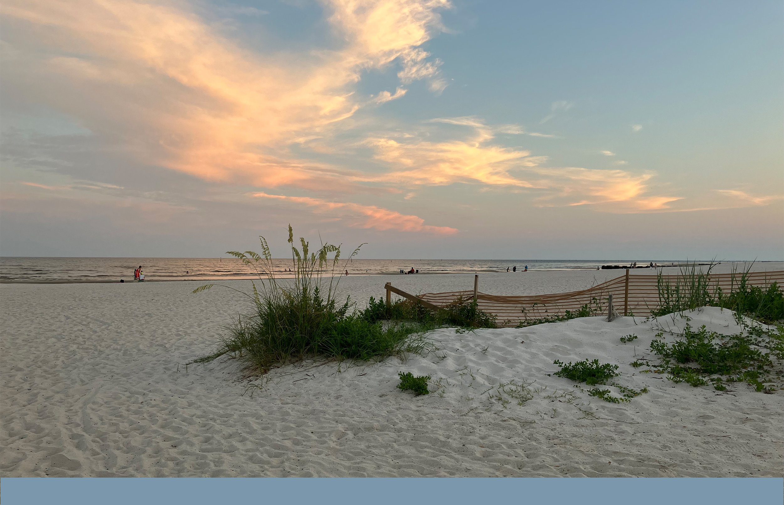 A serene beach scene at sunset with colorful clouds, sandy shore, some beach plants, and a few distant people near the water.