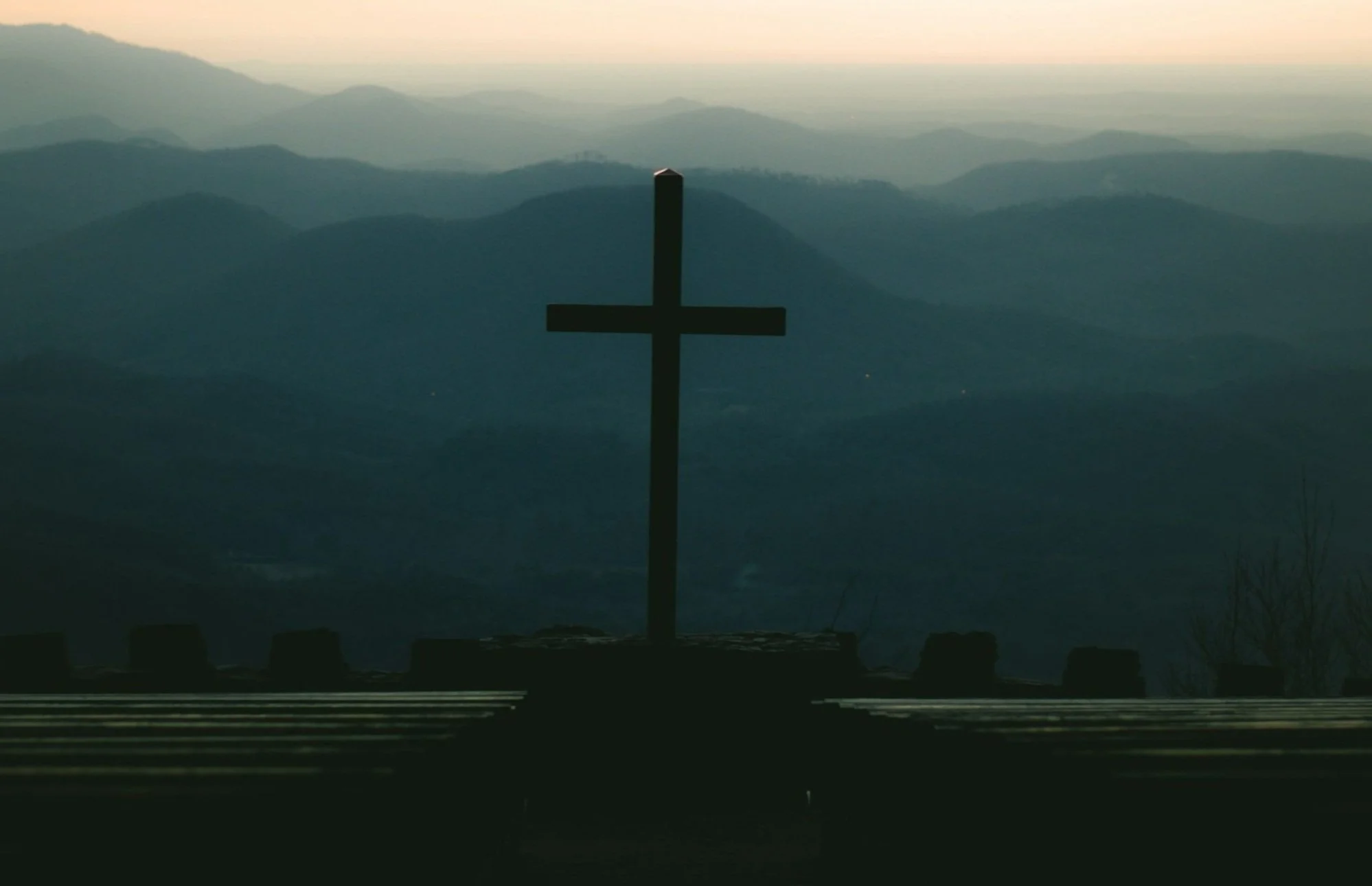 Silhouette of a cross on a hill overlooking a landscape of rolling hills and mountains at dusk or dawn.
