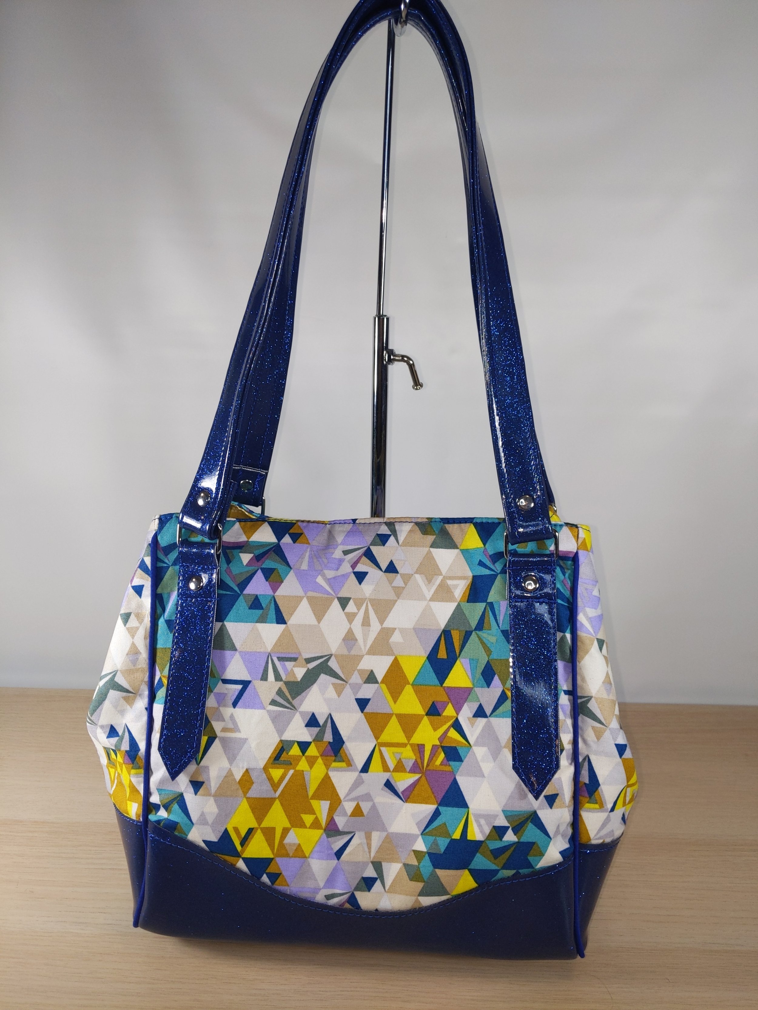 Colorful geometric patterned tote bag with blue shiny straps, placed on a light wooden surface.