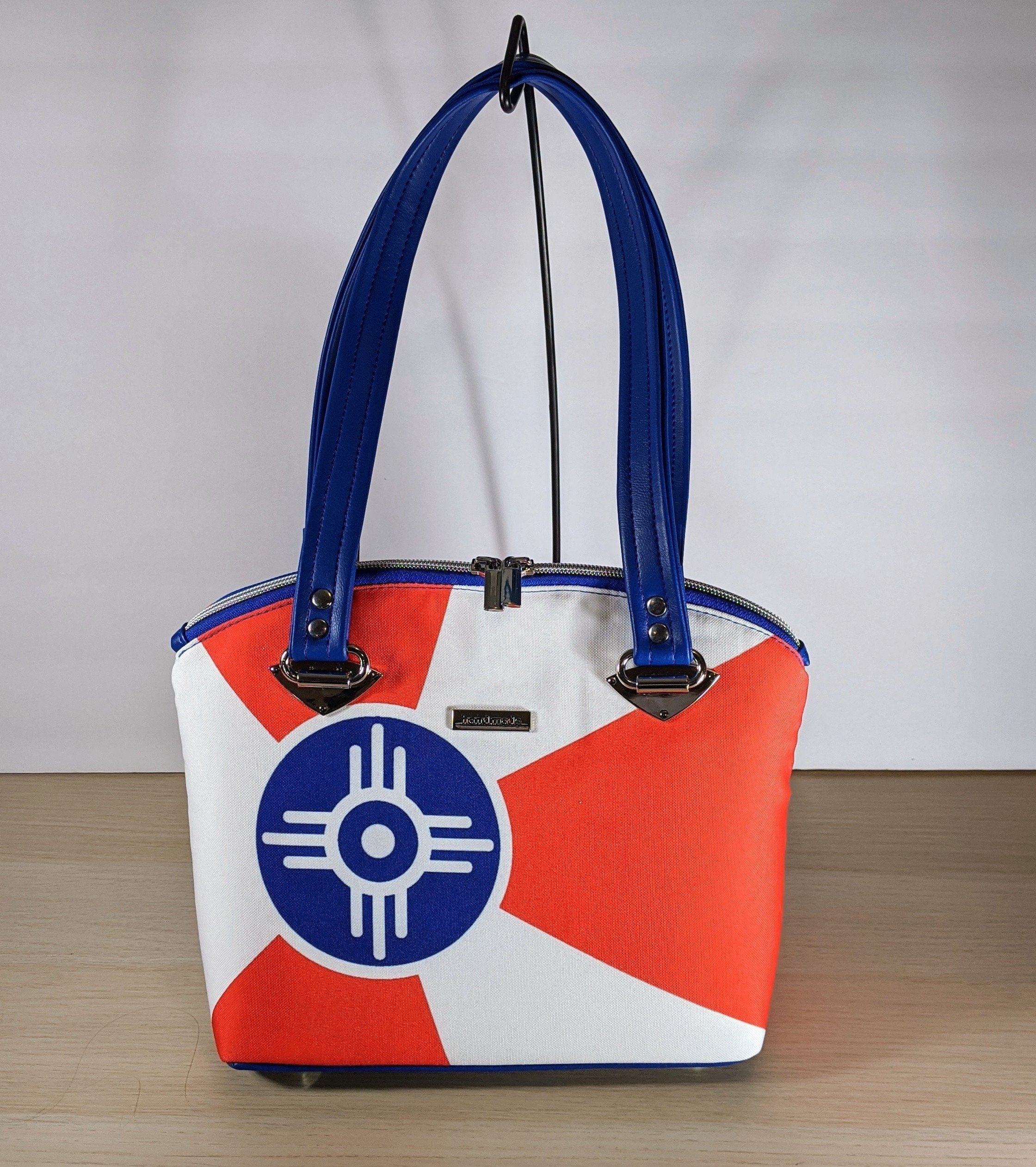 A white handbag with red geometric shapes and a blue circular logo, featuring blue handles and silver hardware, displayed on a light wood surface against a plain background.