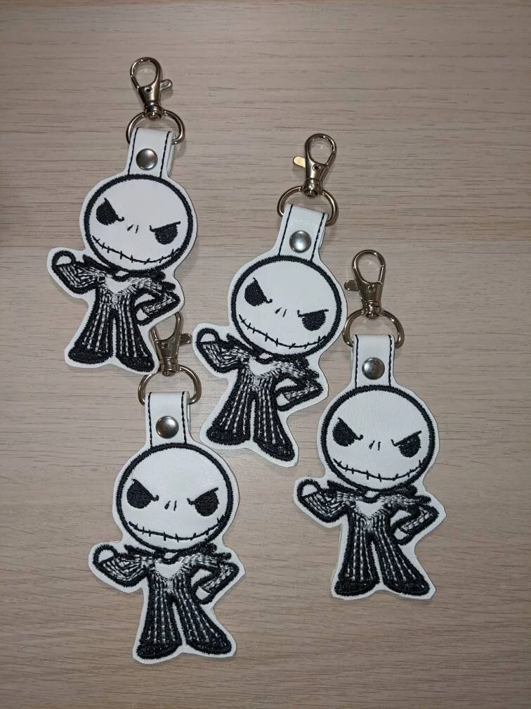 Four embroidered keychains featuring a cartoon character with an evil grin, large black eyes, and dressed in a striped suit, laid on a light wooden surface.