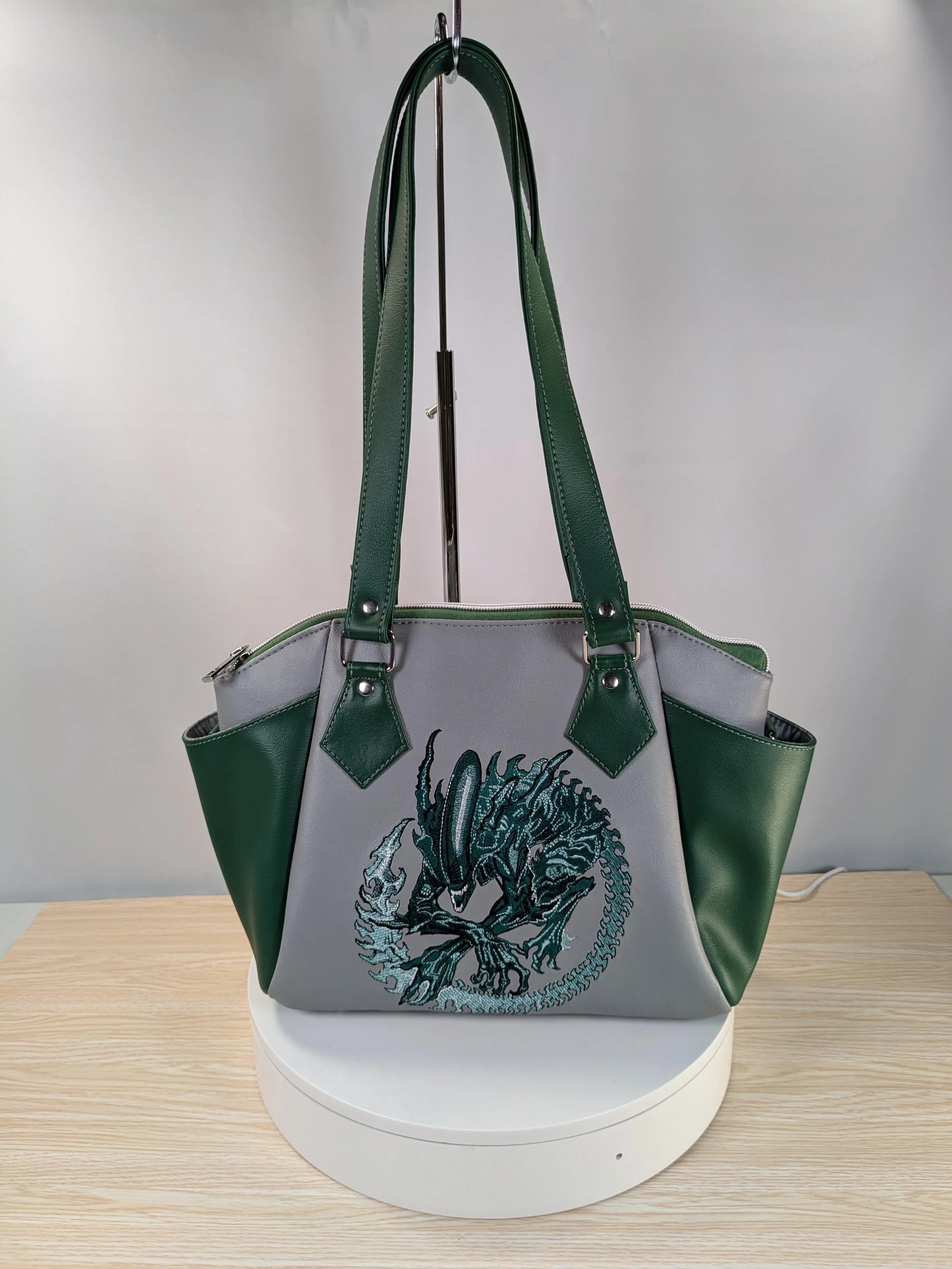 A gray handbag with green straps and side pockets, featuring an embroidered dragon design on the front.