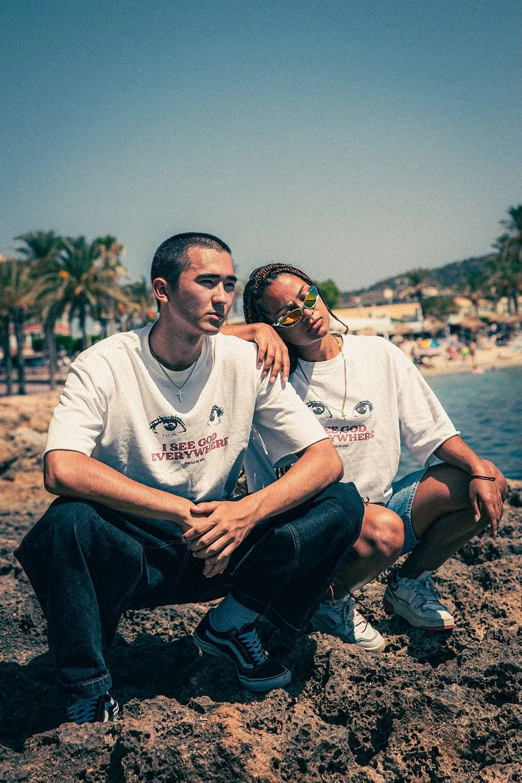 Zwei Jugendliche sitzen auf einem Felsen am Strand bei Sonnenbrille und T-Shirts mit Augenmotiv und Text. Im Hintergrund sind Palmen, Strand und Menschen sichtbar.