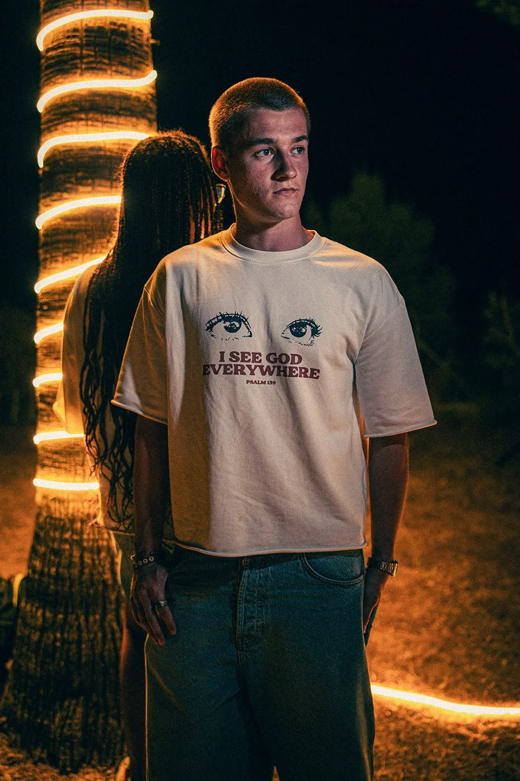 Ein junger Mann mit kurze Haare trägt ein weißes T-Shirt mit Augen und dem Text "I SEE GOD EVERYWHERE" und einer Bibelstelle. Im Hintergrund ist eine Palme mit Lichtern und eine andere Person mit dreadlocks.