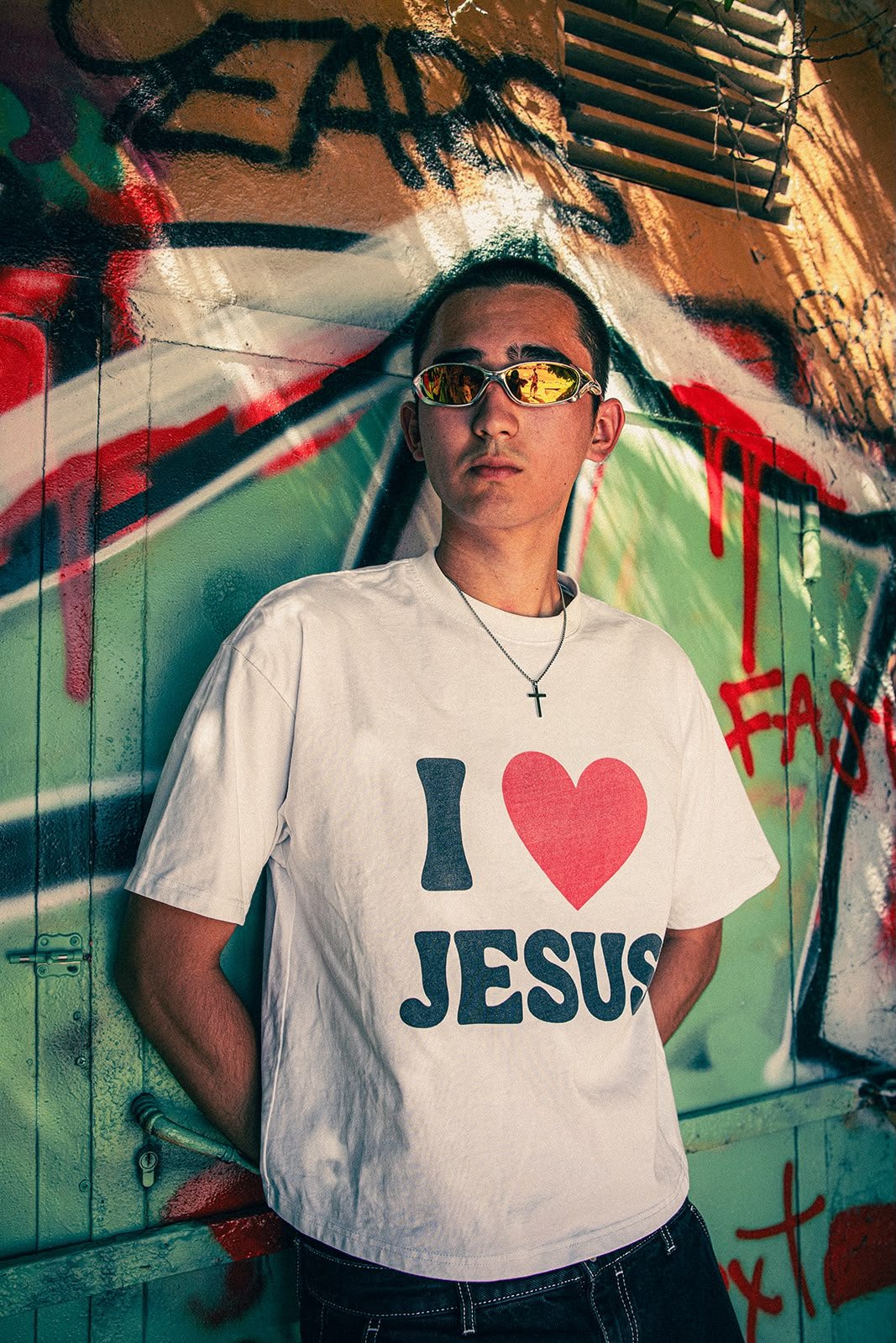 Junge Person mit Sonnenbrille und Kreuzkette, trägt T-Shirt mit der Aufschrift 'I love Jesus', steht vor einer bunten Graffiti-Wand.