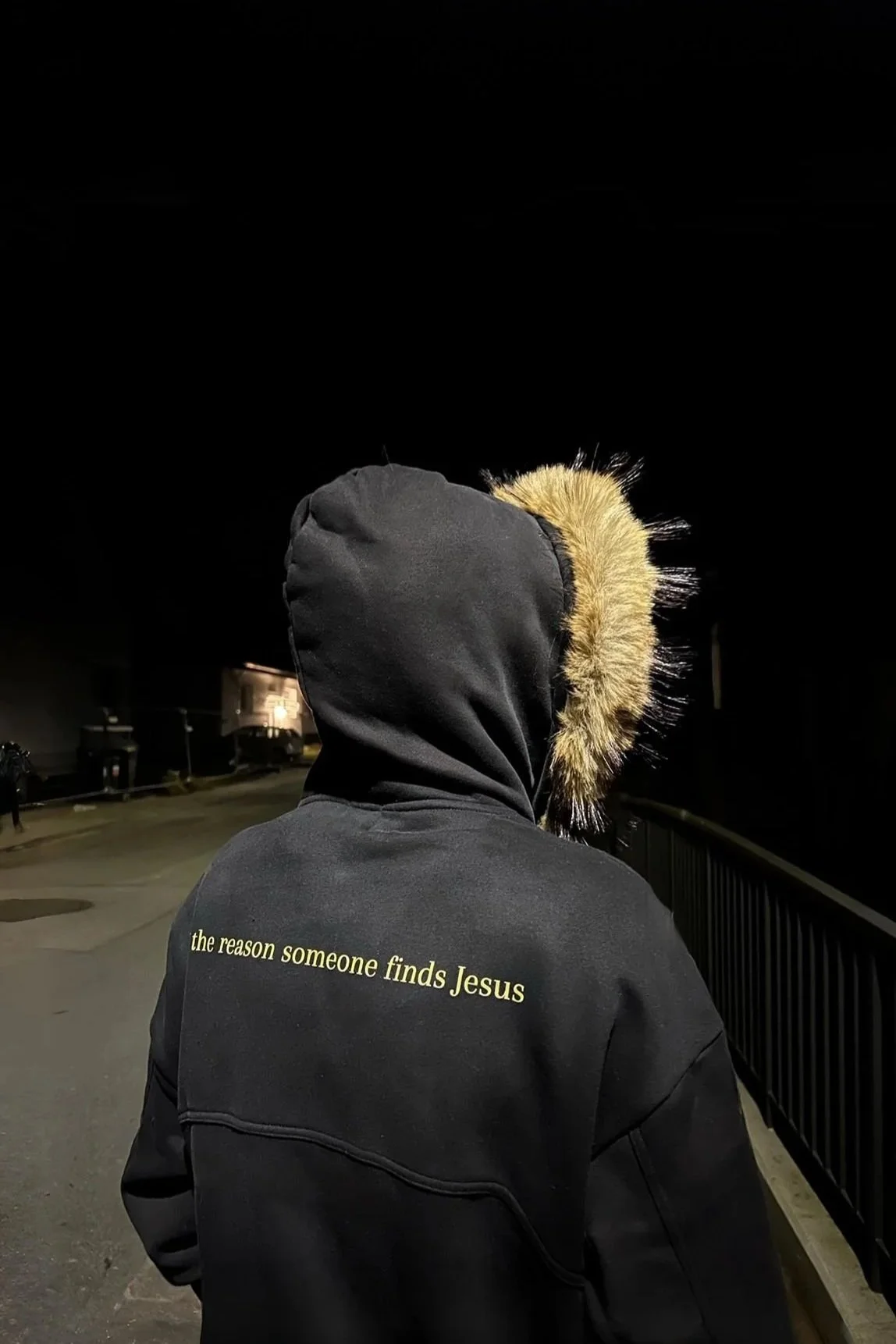 Eine Person trägt eine schwarze Kapuzenjacke mit der Aufschrift "the reason someone finds Jesus" auf dem Rücken, bei Nacht draußen auf einer Brücke.