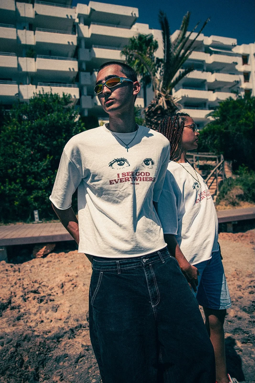 Zwei junge Frauen stehen draußen in der Sonne, tragen weiße T-Shirts mit Aufdruck und sind vor einem modernen Gebäudekomplex mit Balkon. Im Hintergrund sind Bäume und eine Treppe sichtbar.