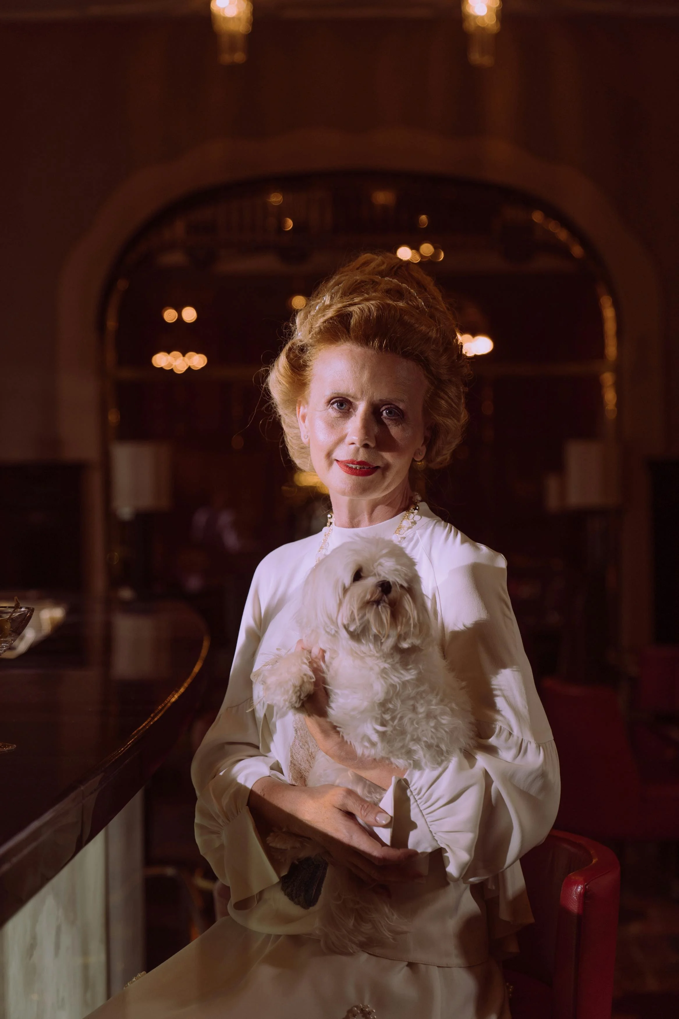 An elderly woman with red hair in an elegant white dress holding a small, fluffy white dog in her arms. She is inside a warmly lit, sophisticated room with dark wood and ornate decor.
