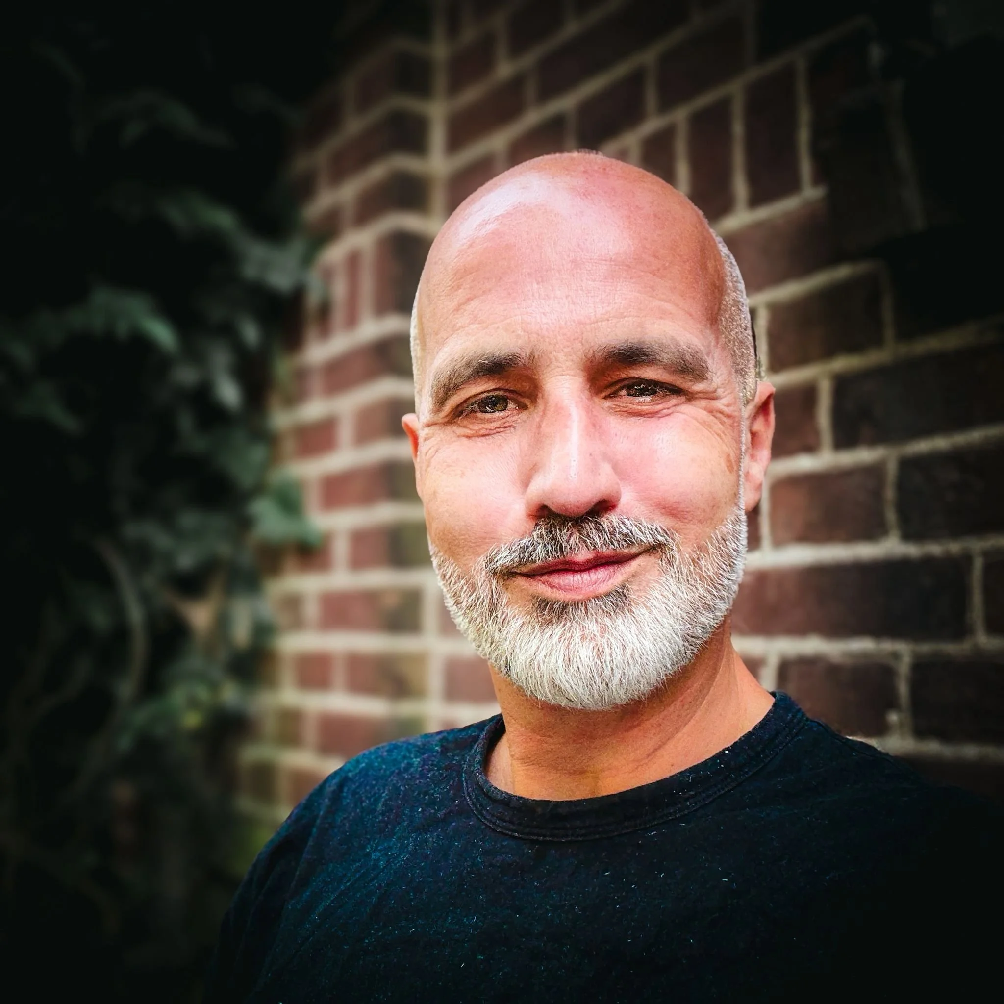 A middle-aged man with a white beard, smiling, standing outdoors in front of a brick wall and greenery.
