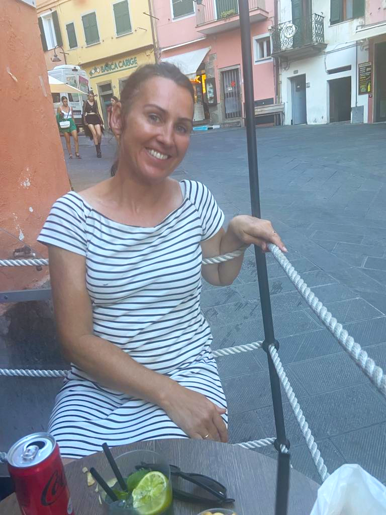 Fornøyd kvinnelig kunde i stripete kjole smiler mens hun sitter utendørs på en terrasse eller gatekafé, med drinker og en åpen boks med Coca-Cola foran seg, omgitt av fargerike bygninger.
