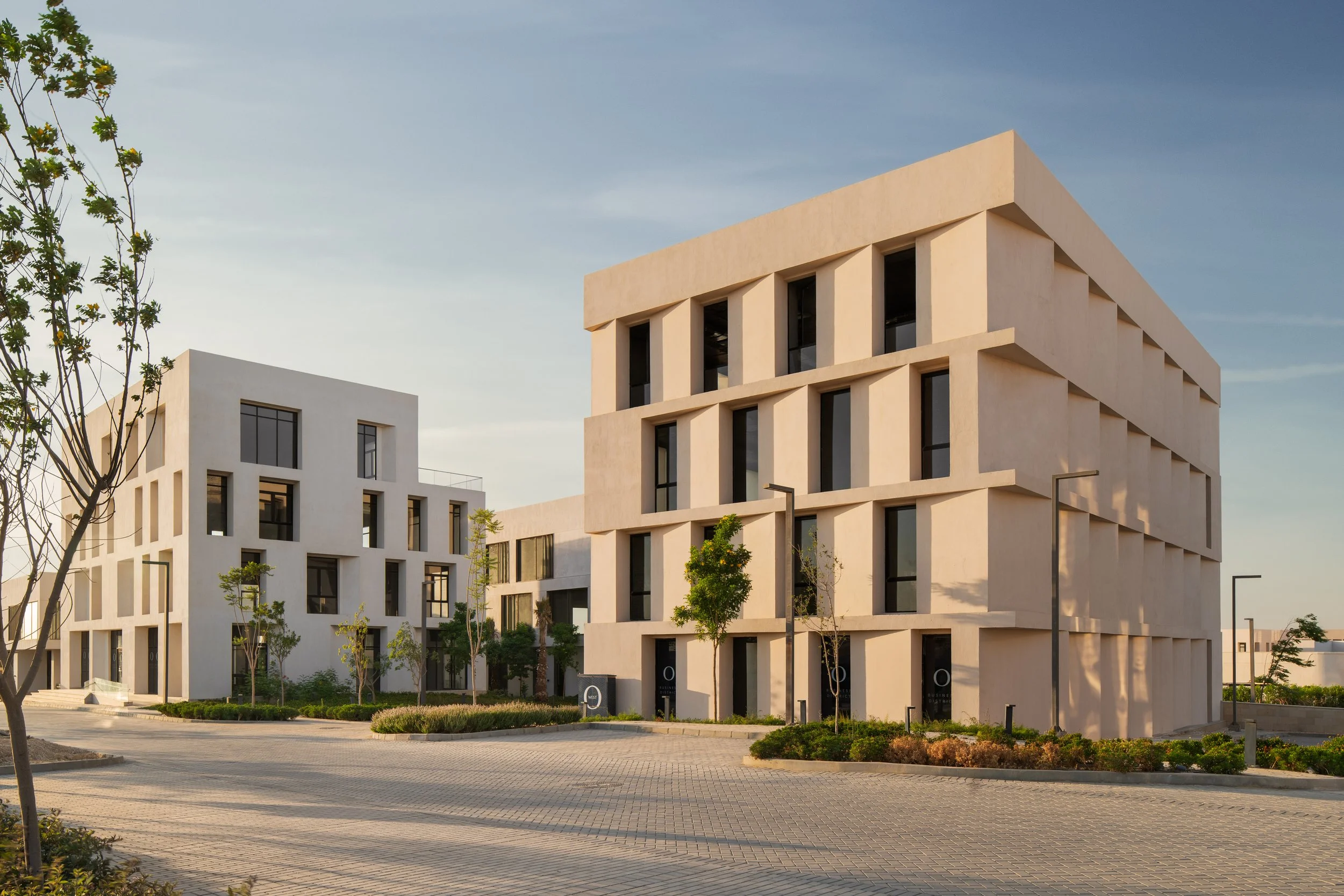 Modern residential buildings with geometric design and large windows, surrounded by landscaped greenery and paved walking paths, under a clear sky.