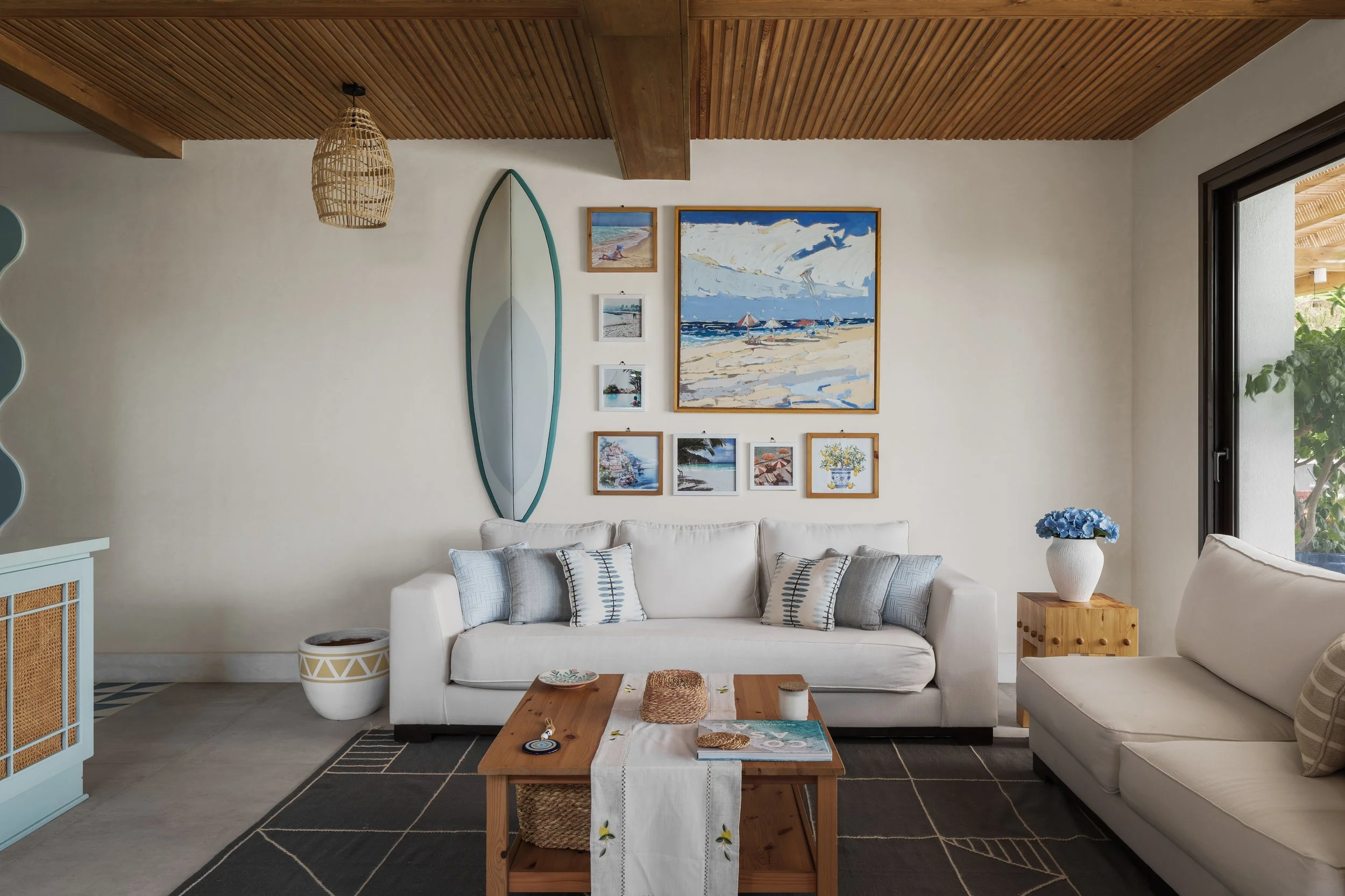 Beach-themed living room with white sofas, a wooden coffee table, framed artworks of beaches and ocean scenes, a surfboard mirror, and a sliding glass door to an outdoor patio.