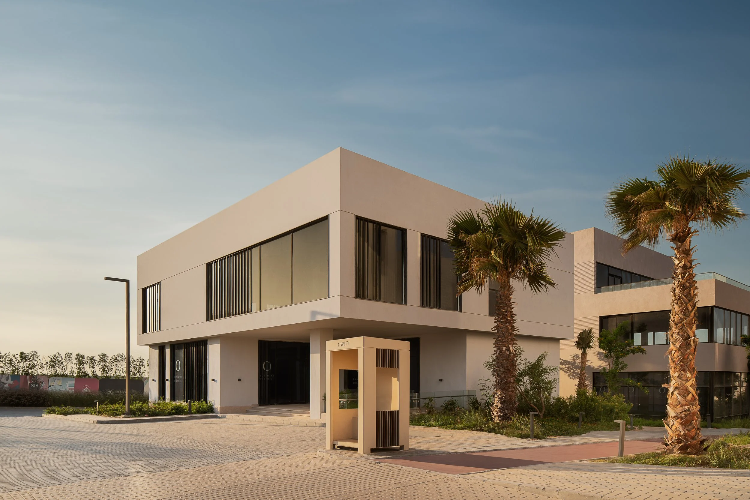 Modern two-story building with large windows, surrounded by palm trees and a paved courtyard, under a blue sky.