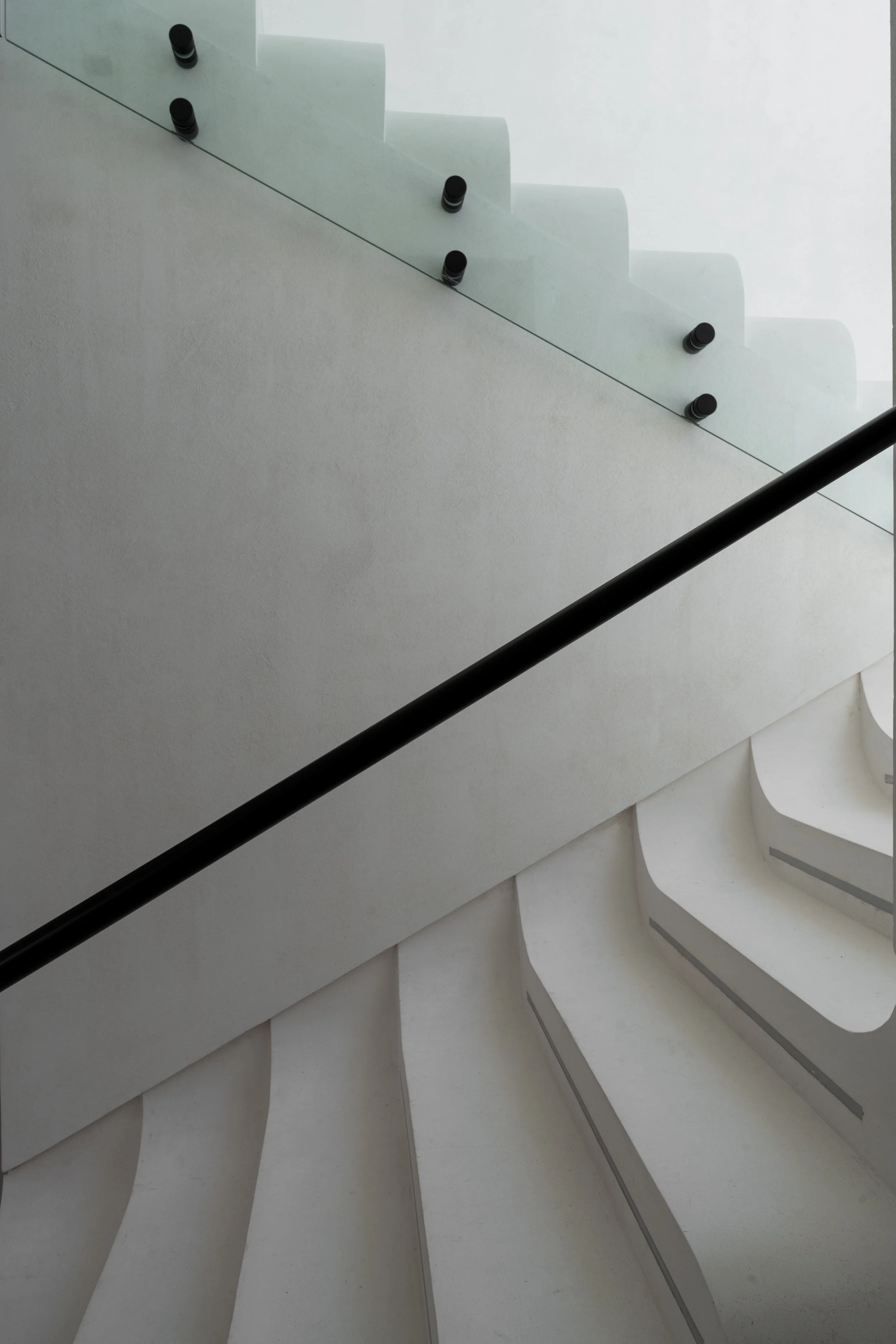 Abstract interior architectural view with white curved stairs, a glass railing, and a black handrail. The perspective looks up the staircase.