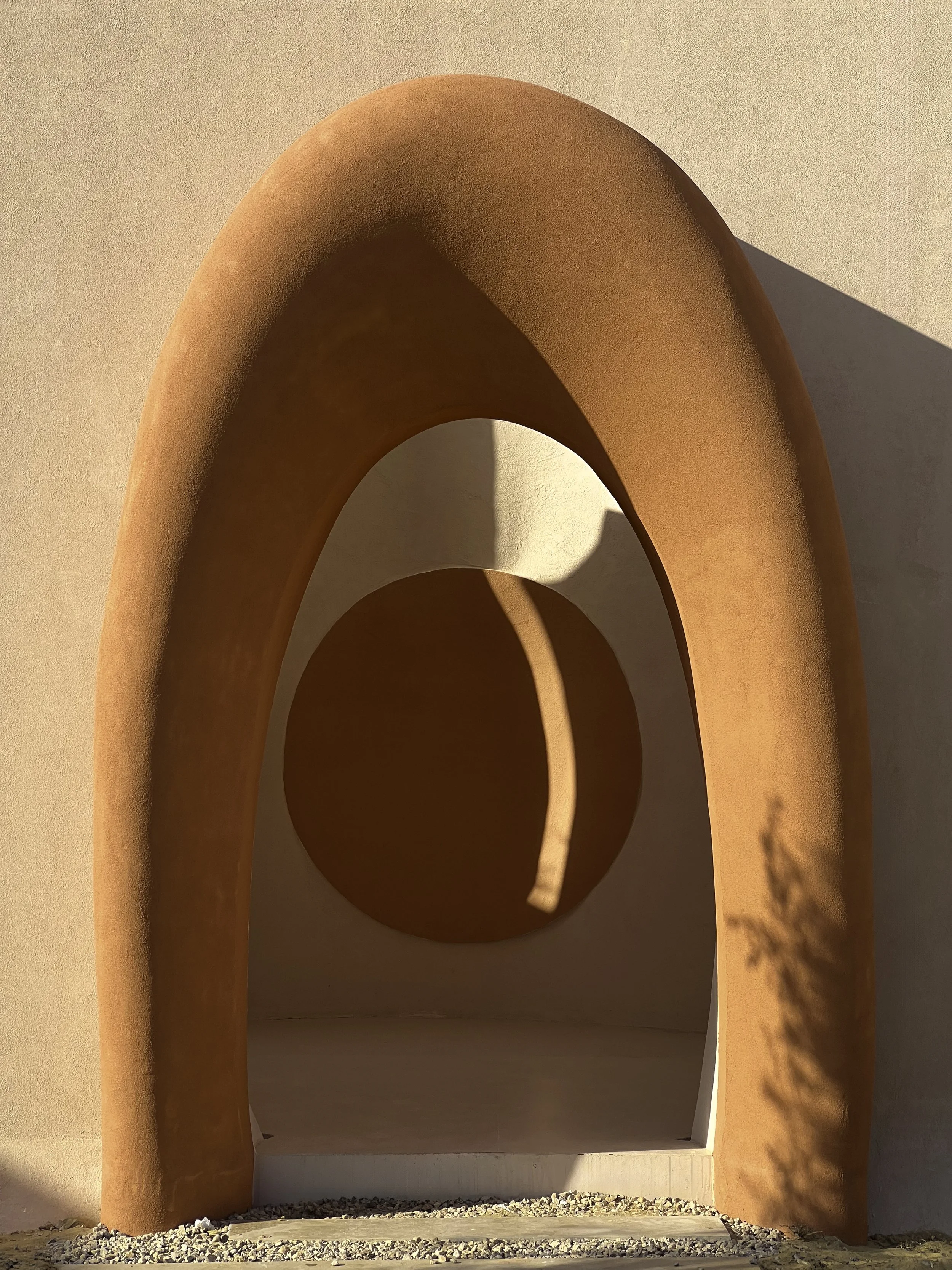 A minimalist architectural structure with a terracotta-colored arch and a circular opening, casting a shadow on a beige wall.