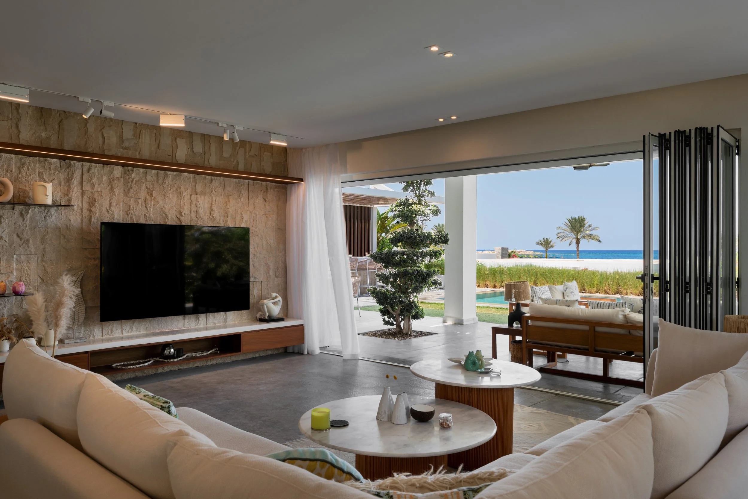 Living room with large open window showing a view of the ocean, palm trees, outdoor seating, and a garden, featuring a beige sofa, marble coffee tables, a stone wall with a mounted flat-screen TV, and decorative objects.