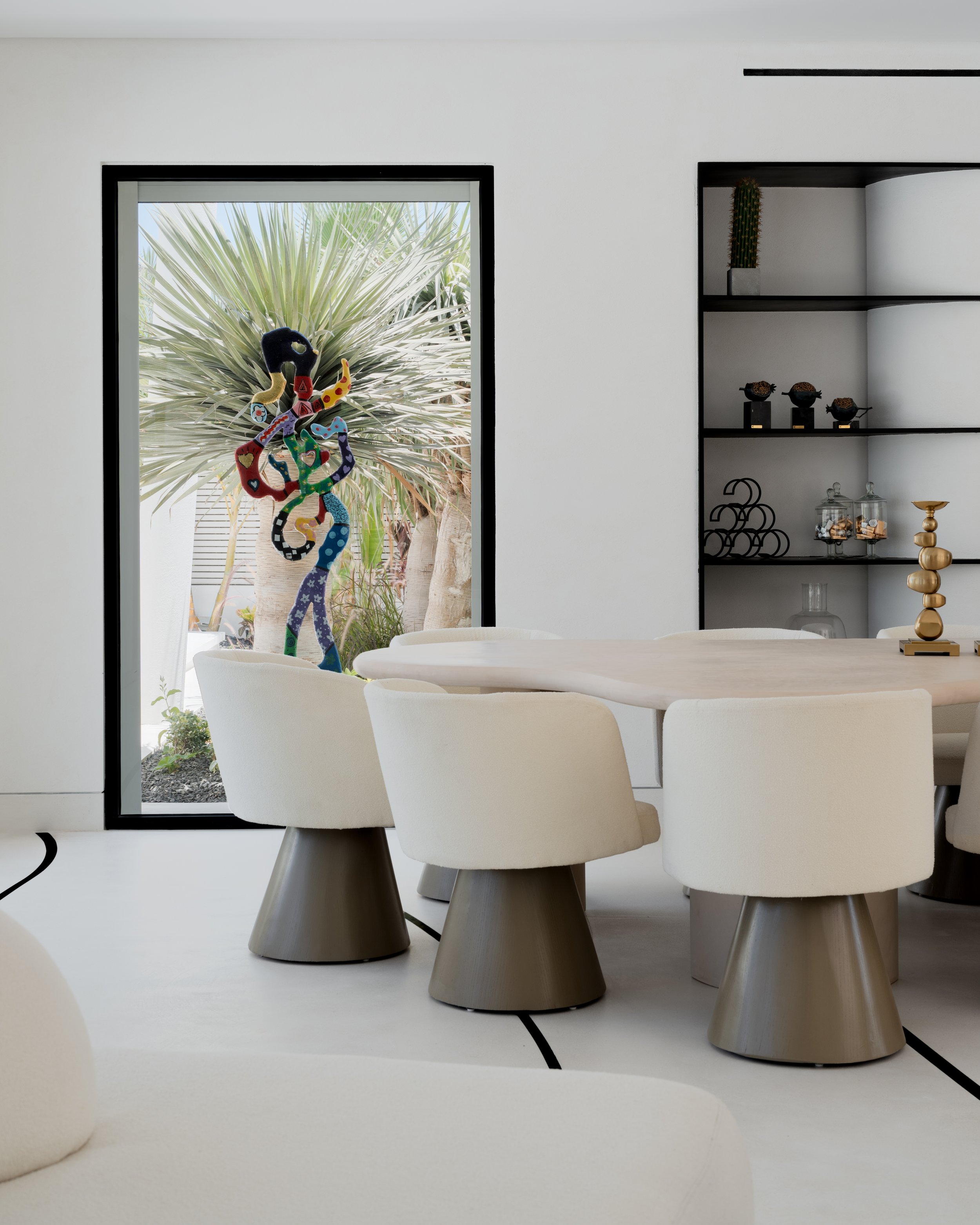 Modern dining room with white chairs, a wooden table, and decorative shelving. Outside window shows a palm tree with a colorful sculpture resembling a snake or dragon.