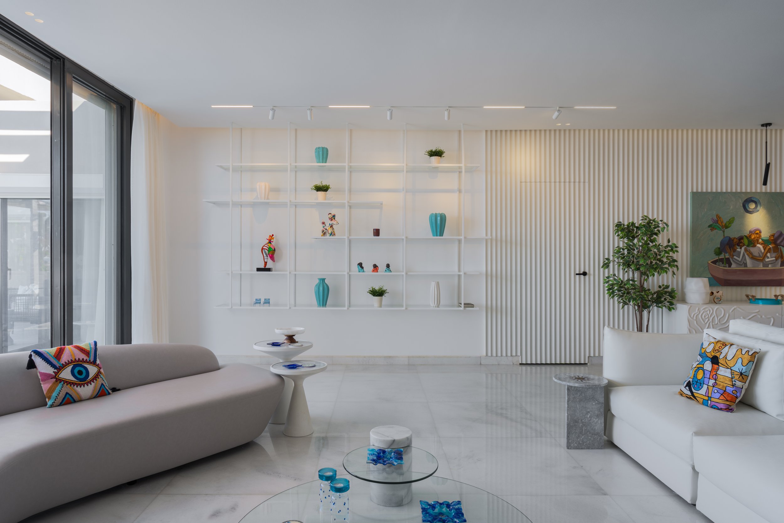 Modern living room with white sofas, decorative pillows, glass tables, and a white wall with shelves holding vases and small decorative objects. Large glass sliding door on the left, plant near the right sofa, abstract painting on the right wall.