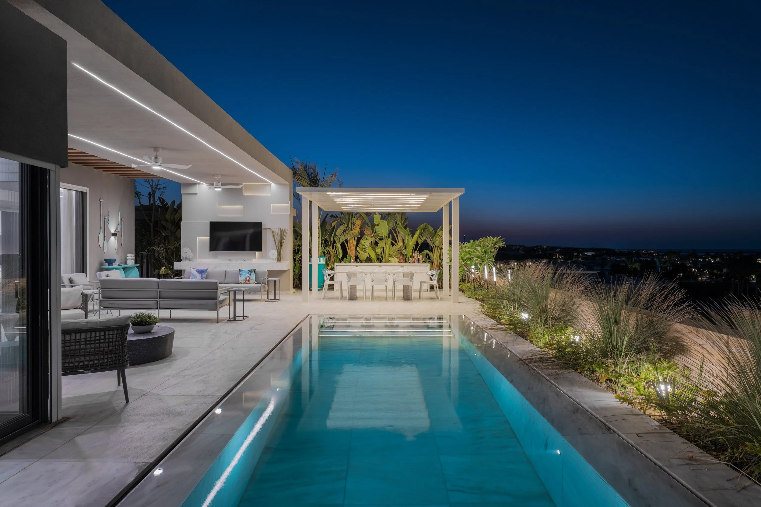 Modern backyard patio at night with a swimming pool, outdoor seating, dining area under a pergola, and decorative lighting, overlooking a cityscape.