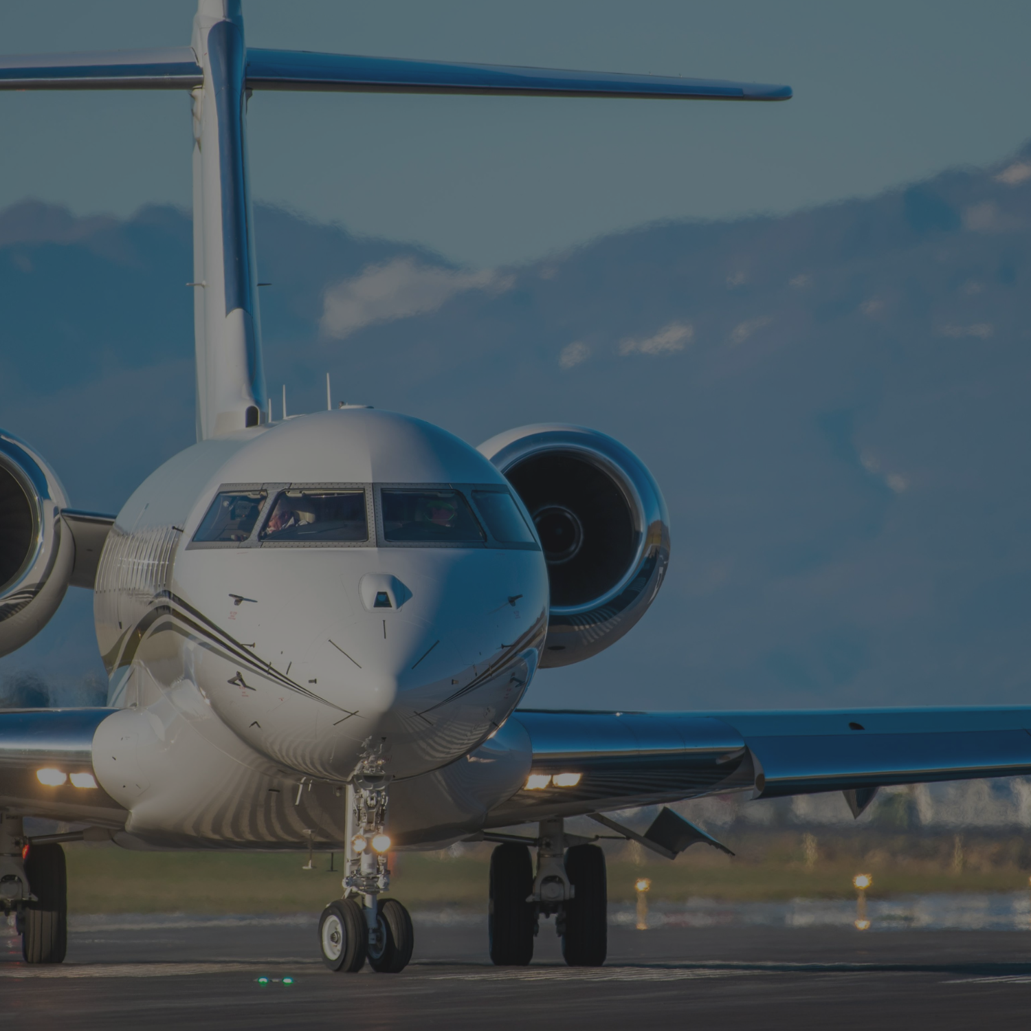 Front view of a white private jet on the runway, with mountains and a clear sky in the background.