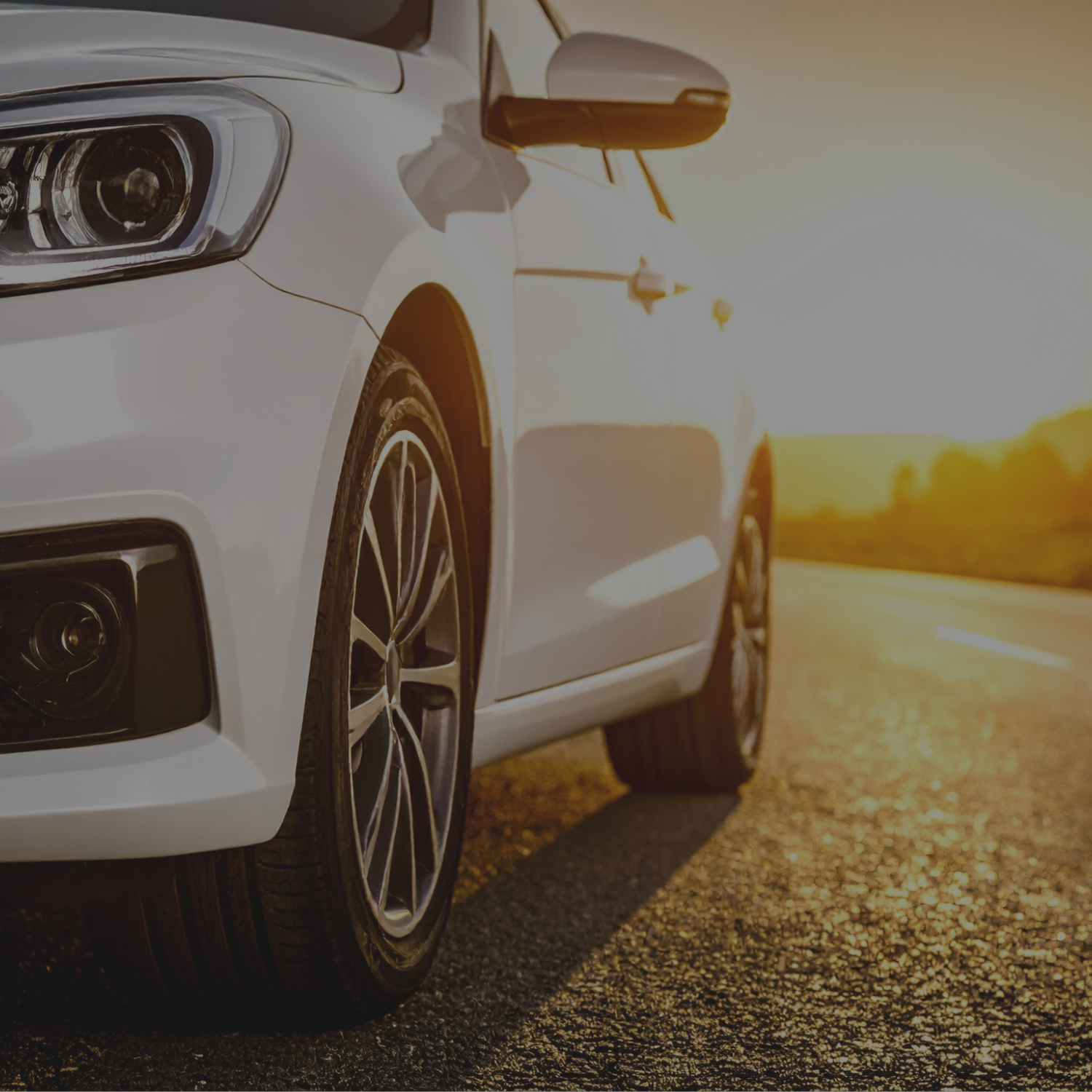 Close-up of a white car parked on the side of a road during sunset, with focus on the front wheel and headlight.