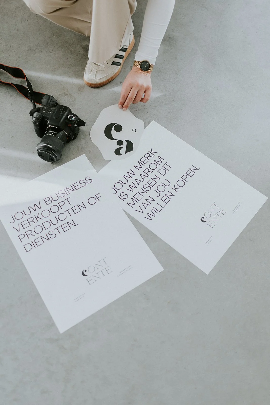 Persoon die een logo op papier en twee posters op de grond plaatst, naast een digitale camera en een horloge. De posters bevatten teksten in het Nederlands over het verkopen en de marketing van producten of diensten.