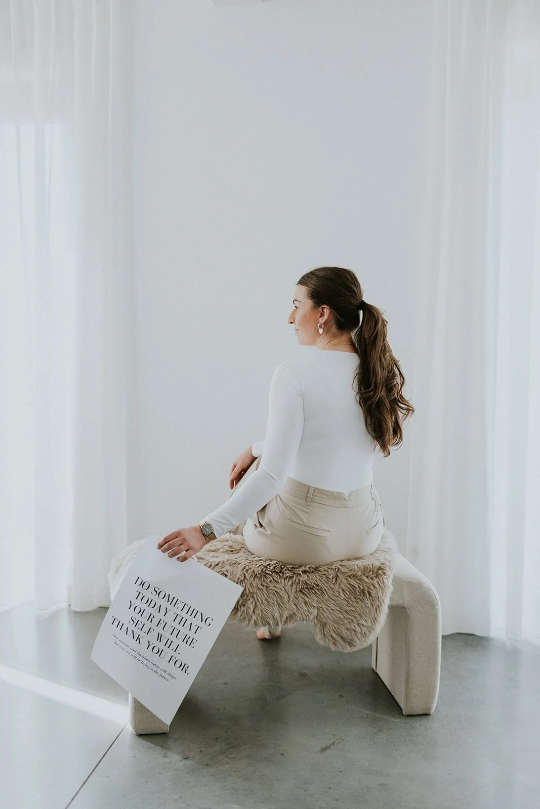 Vrouw zit op een bank met een schapenvacht, vastgelegd in een lichte kamer met witte gordijnen. Ze heeft bruin haar in een paardenstaart en draagt een witte top en beige broek. Ze houdt een bord met een motiverende quote: 'Doe iets vandaag dat je toekomst zelf zal bedanken voor'.