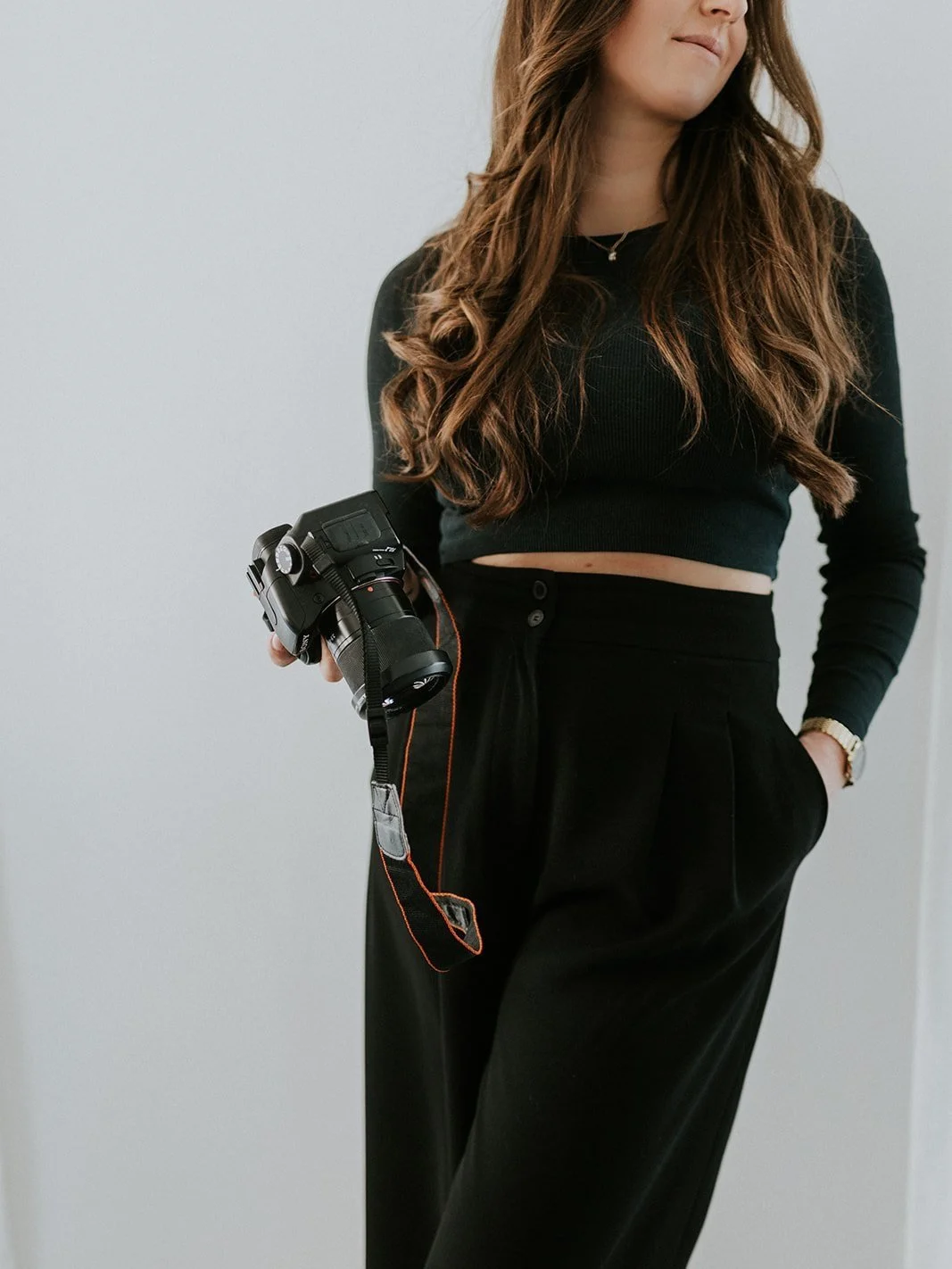 Vrouw in zwart outfit met camera in hand, poseert voor een witte muur. Beeld straalt artistiek gevoel uit.