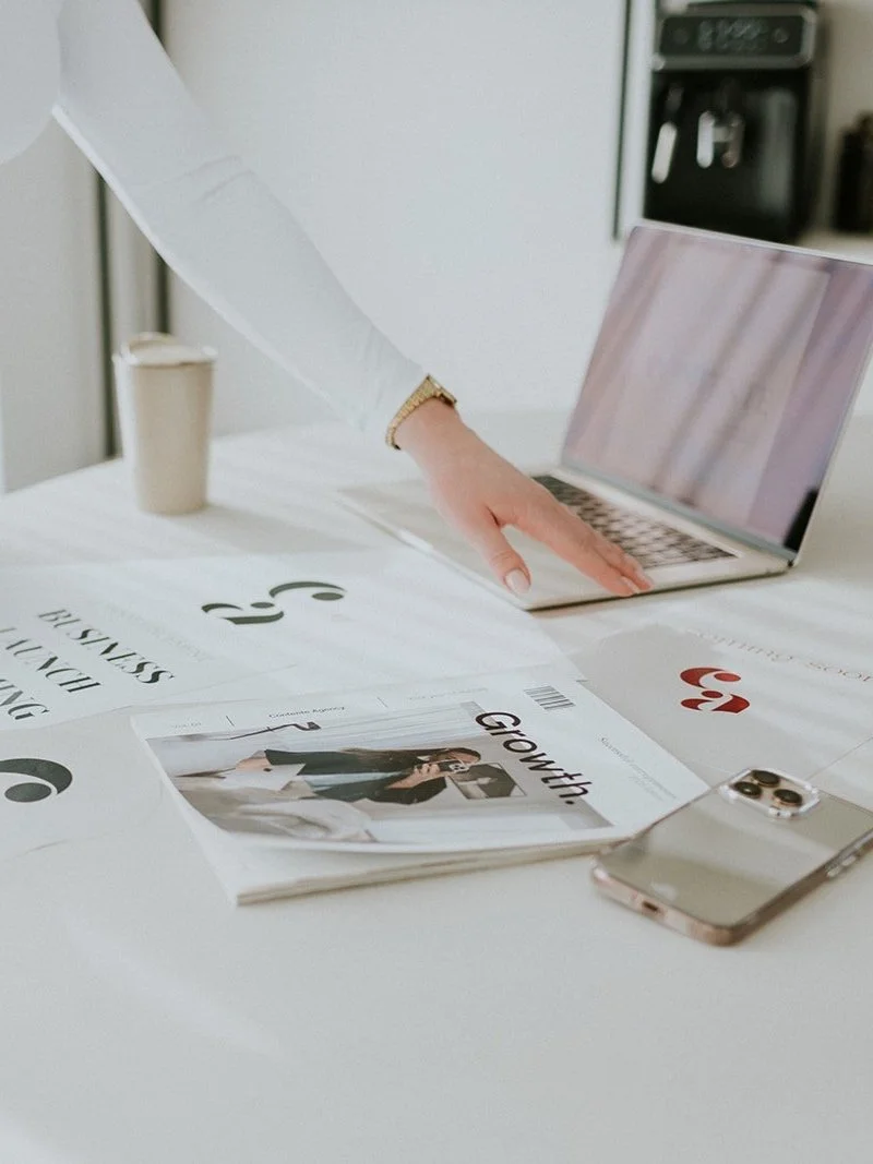 Persoon die met de hand op een open laptop wijst op een bureau met tijdschriften, papieren en een smartphone. Deze vrouw is een moodboard aan het toelichten.