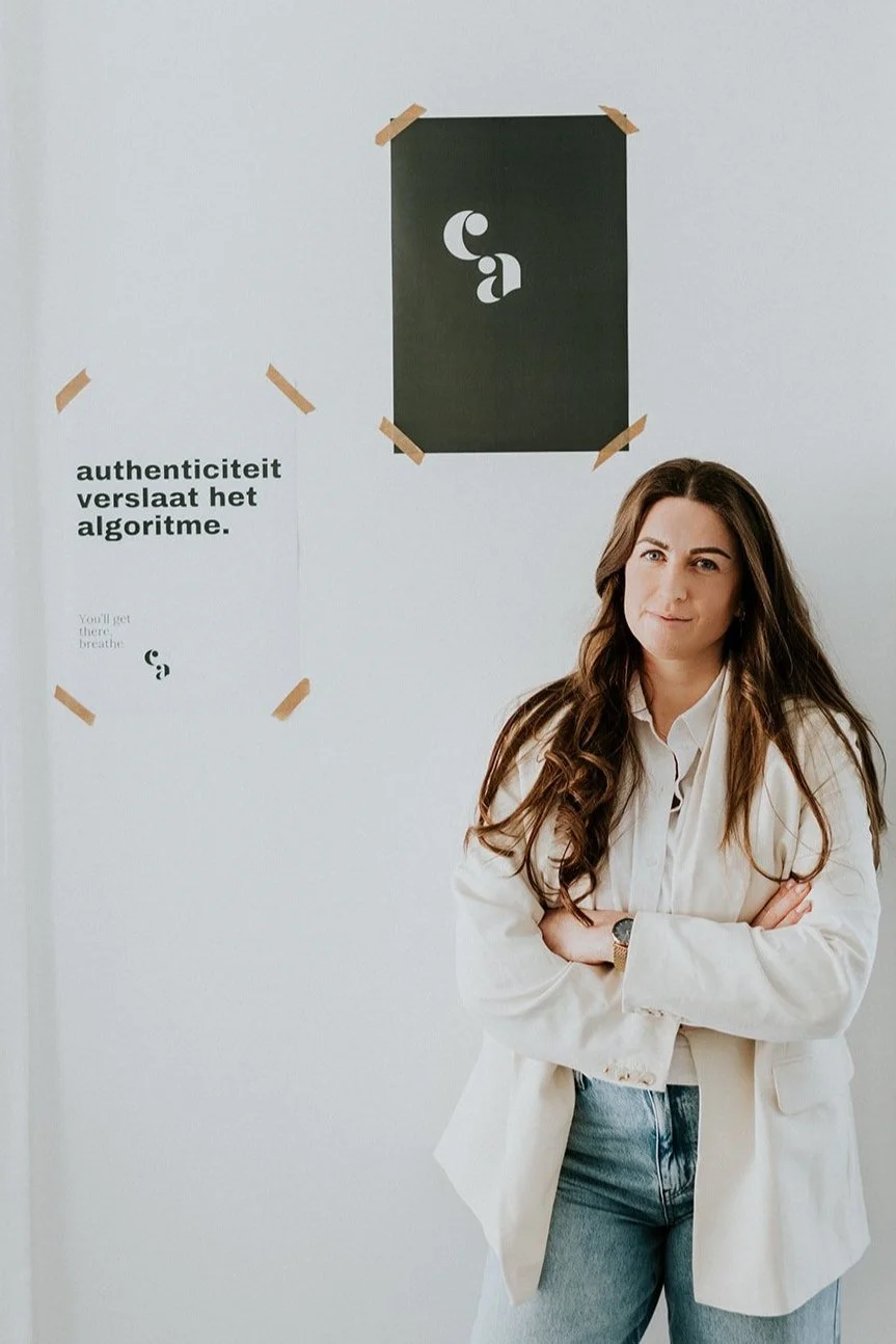 Vrouw met lang, bruin haar in een witte blazerjas staat met gekruiste armen voor een witte muur met twee posters en een grafiek. Eén poster zegt 'authenticiteit verslaat het algoritme' en de andere bevat het abstract logo van haar bedrijf.