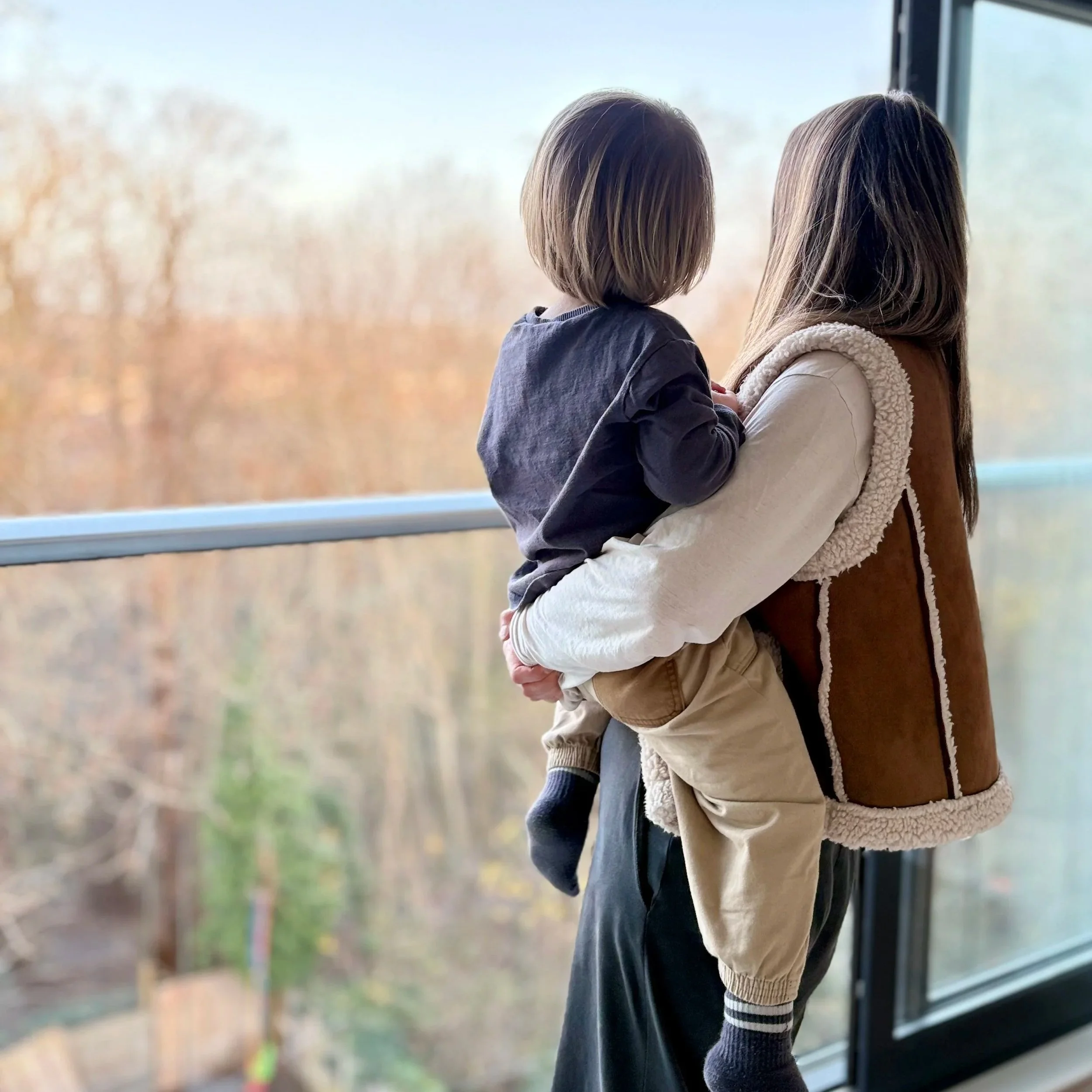 A woman holding a young child while looking out of a large window with a view of trees in autumn.