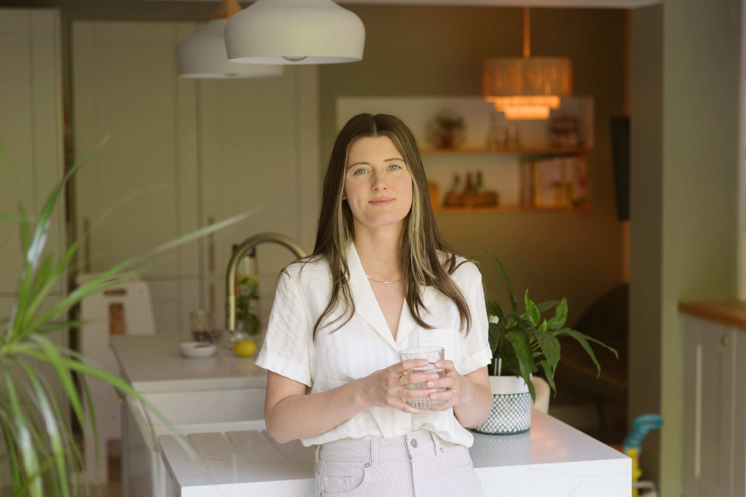 A woman with long brown hair, wearing a white shirt, holding a glass of water in a modern kitchen with plants and warm lighting.