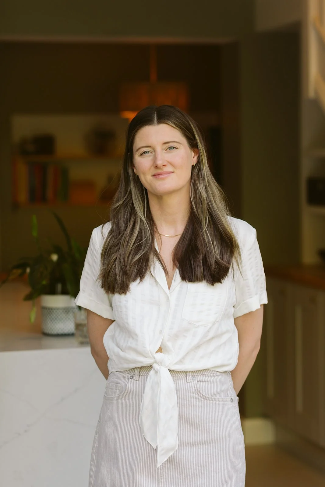 A woman with long brown hair, wearing a white tied-up blouse, standing in a modern kitchen with warm lighting and a plant in the background.