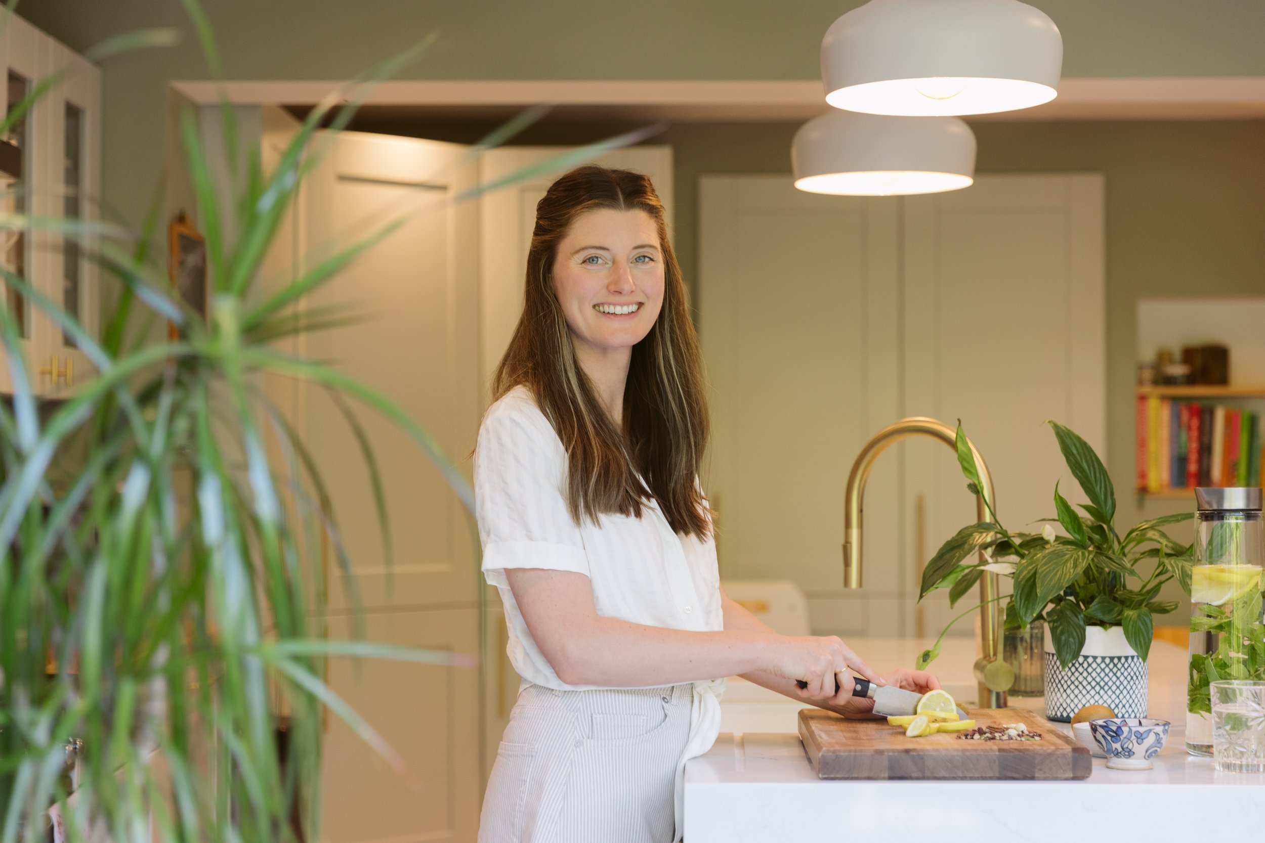 A woman with long brown hair smiling while chopping lemon on a wooden cutting board in a modern kitchen with green walls and plants.