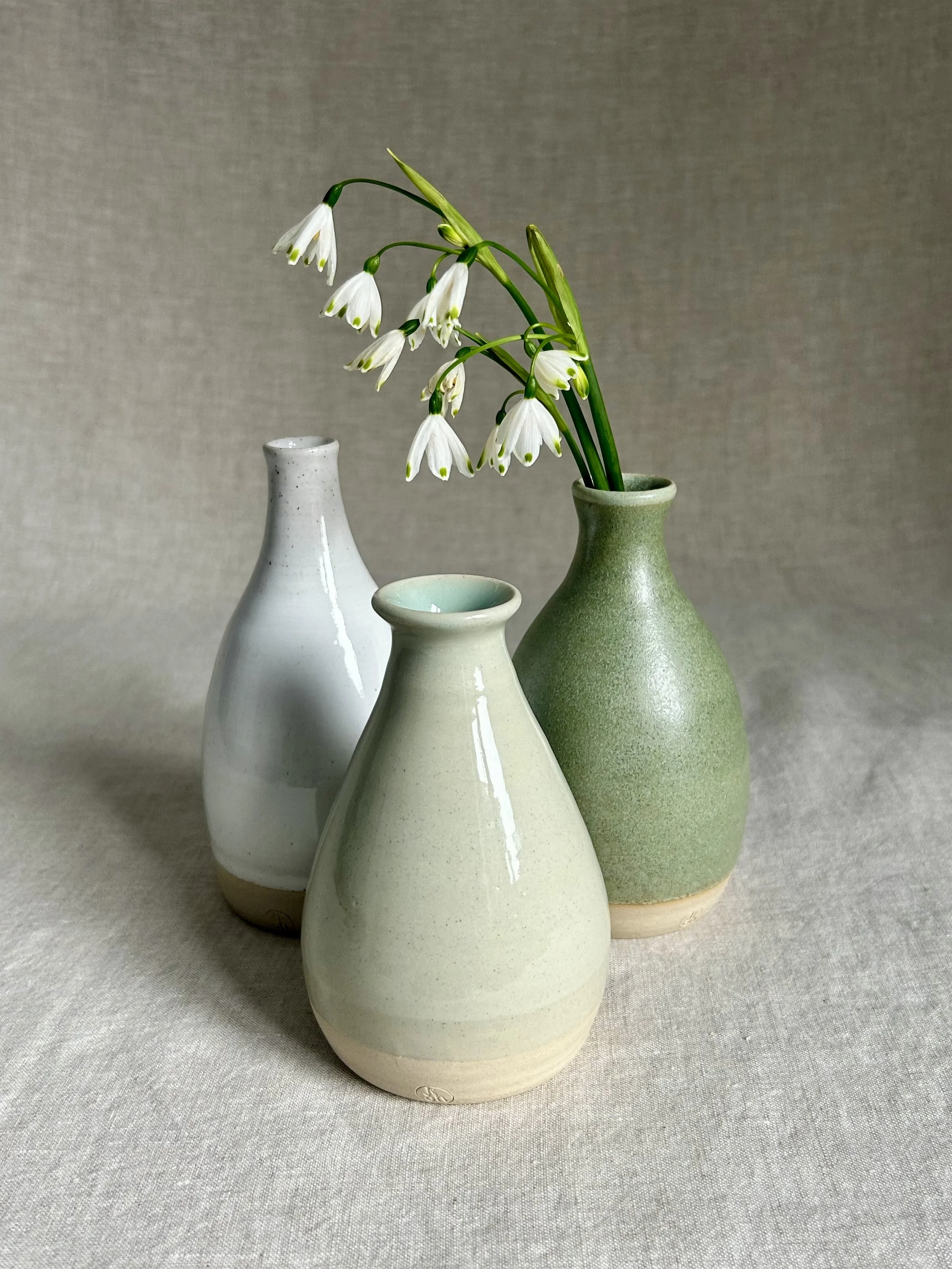 Three ceramic vases of varying shapes and shades of white and green, with one vase holding white snowdrop flowers, placed on a neutral fabric background.
