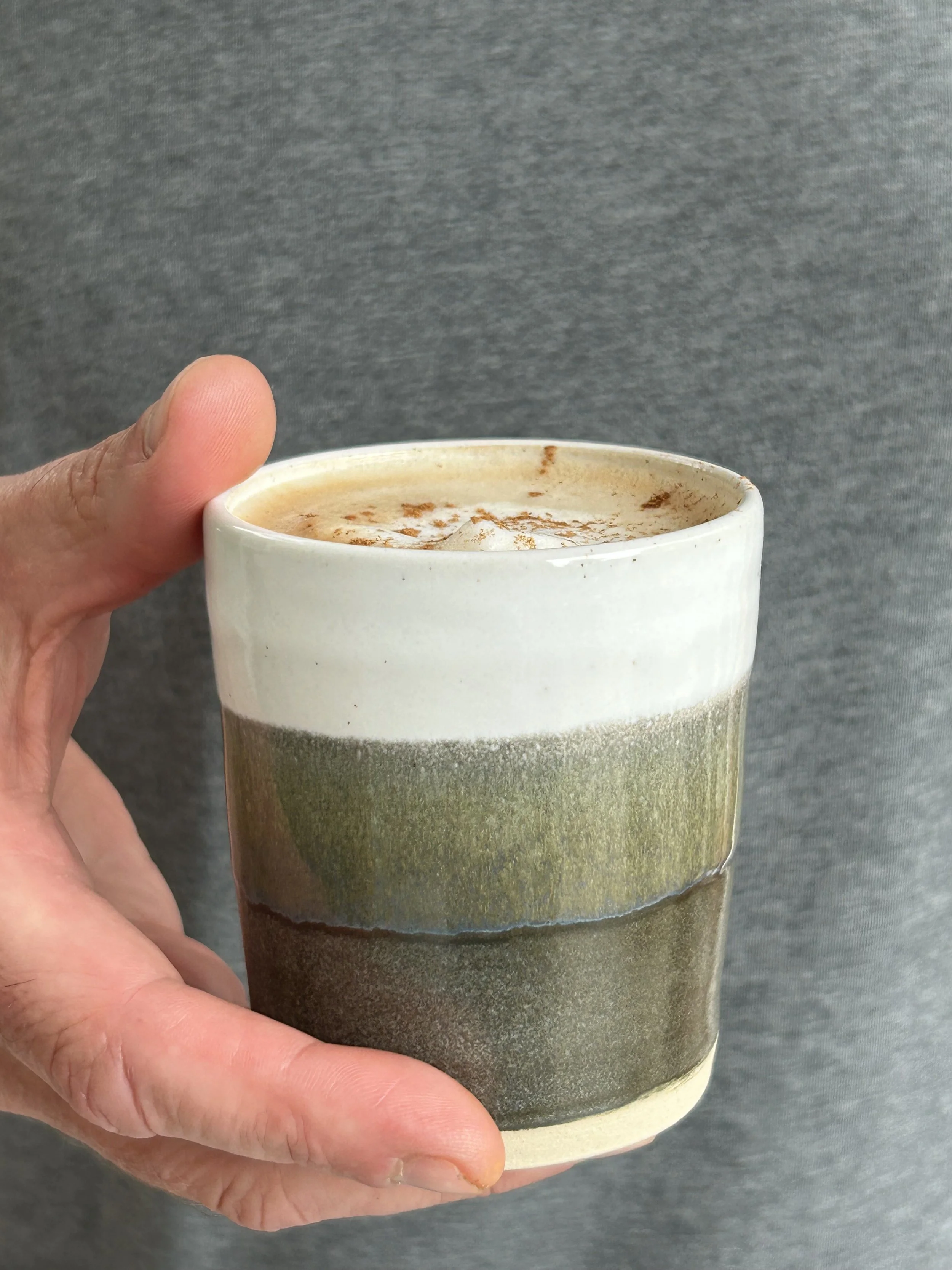 A person's hand holding a handmade coffee cup with a white, grey and green glaze, containing a frothy coffee topped with a dusting of cinnamon.