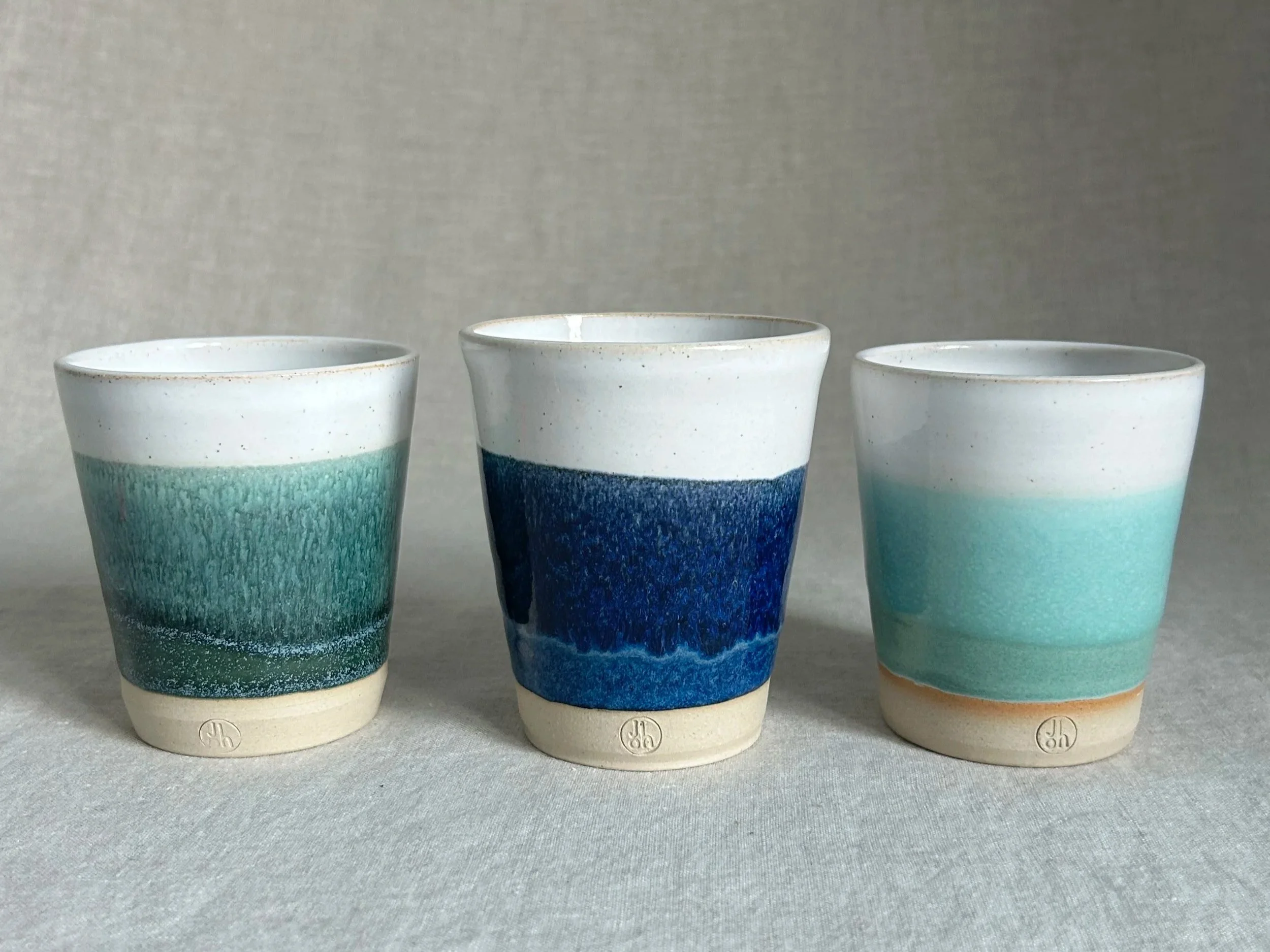 Three ceramic cups with a color gradient from beige at the bottom to teal and blue at the top, set against a plain gray background.