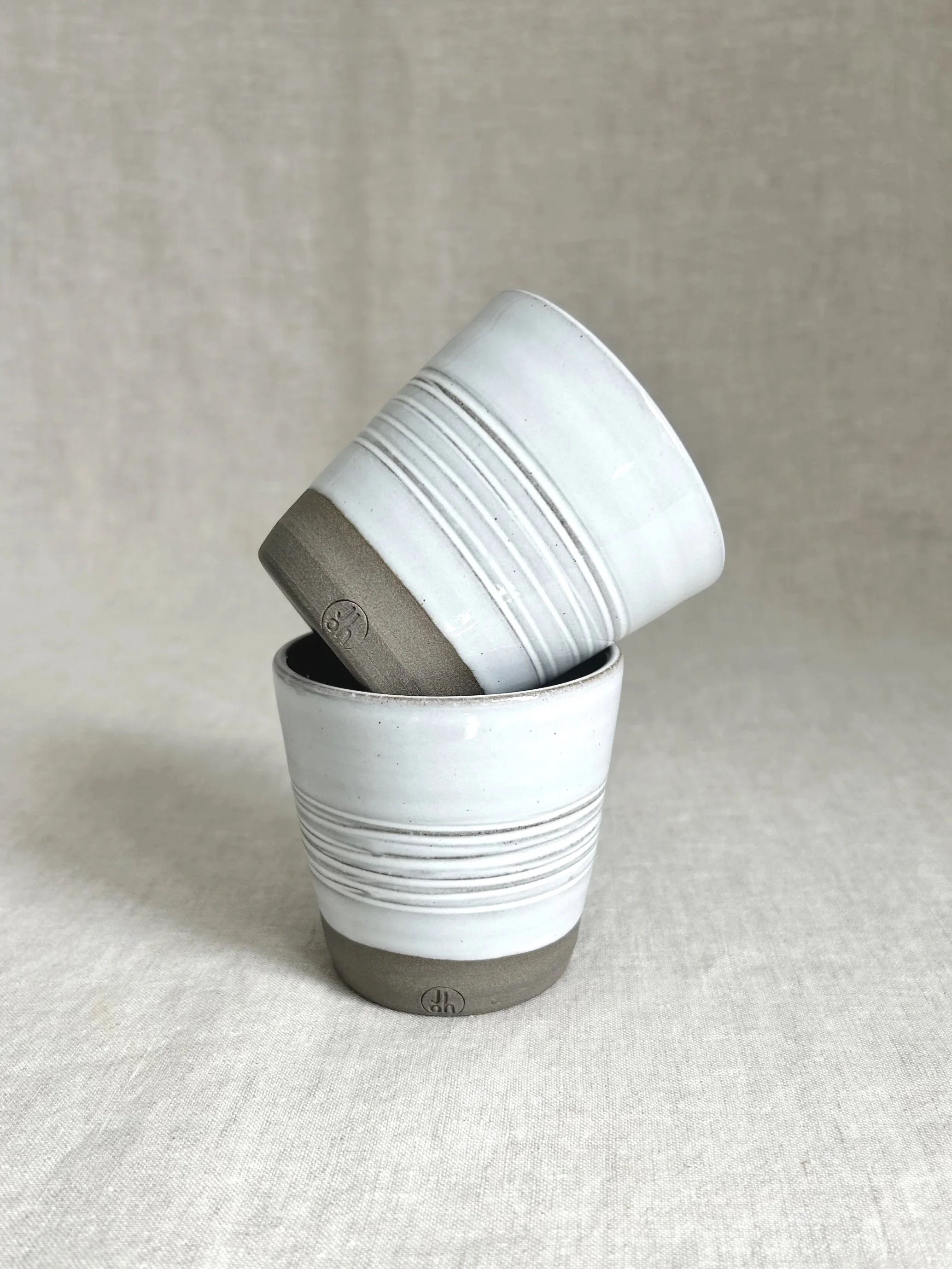 Two stacked cups in chocolate coloured stoneware with a glossy white glaze, one tipped into the other, on a neutral fabric background.