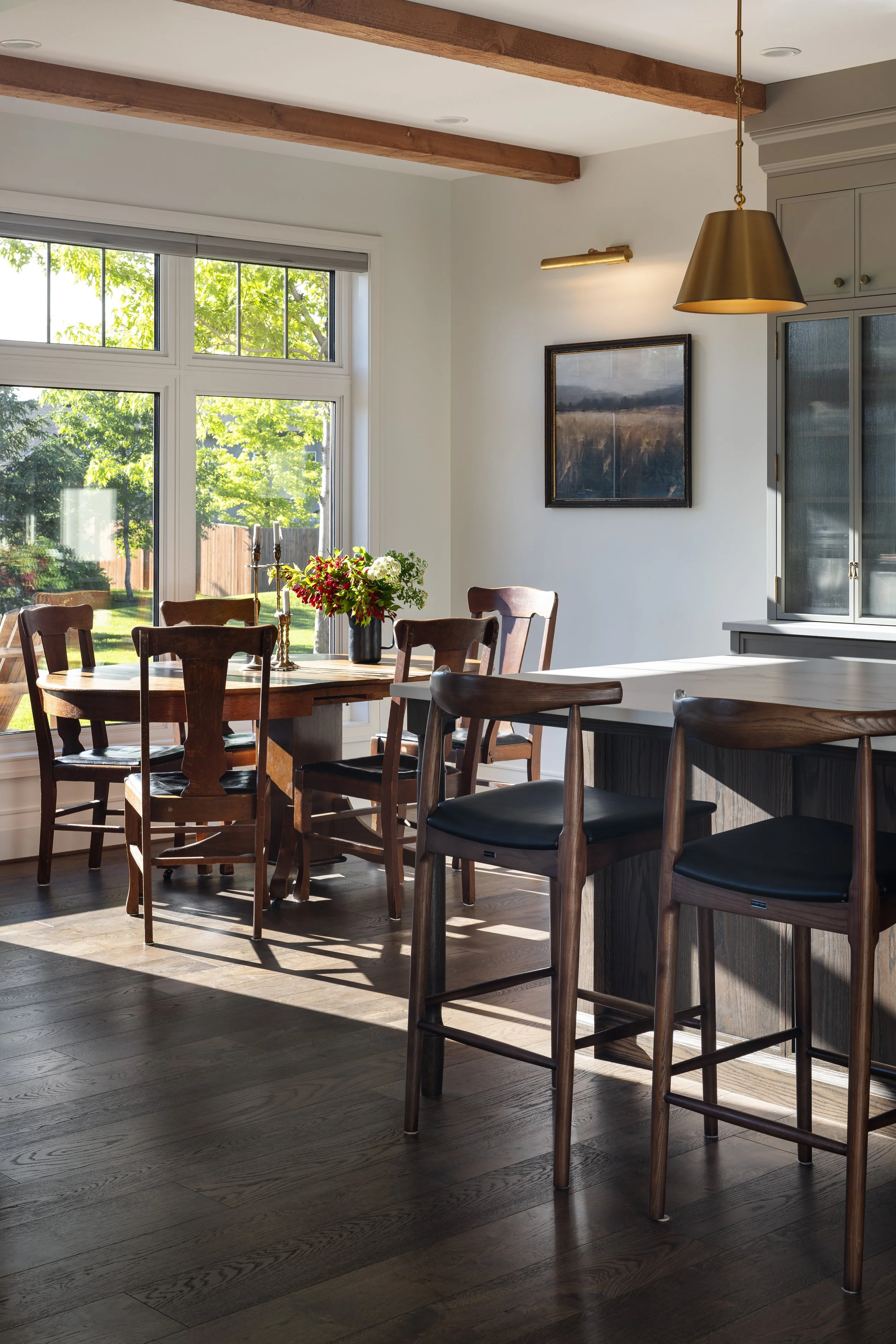 A dining area with a wooden table and six chairs, a large window with a view of a backyard with trees, and a kitchen counter with bar stools. Sunlight streams through the window, illuminating the room.