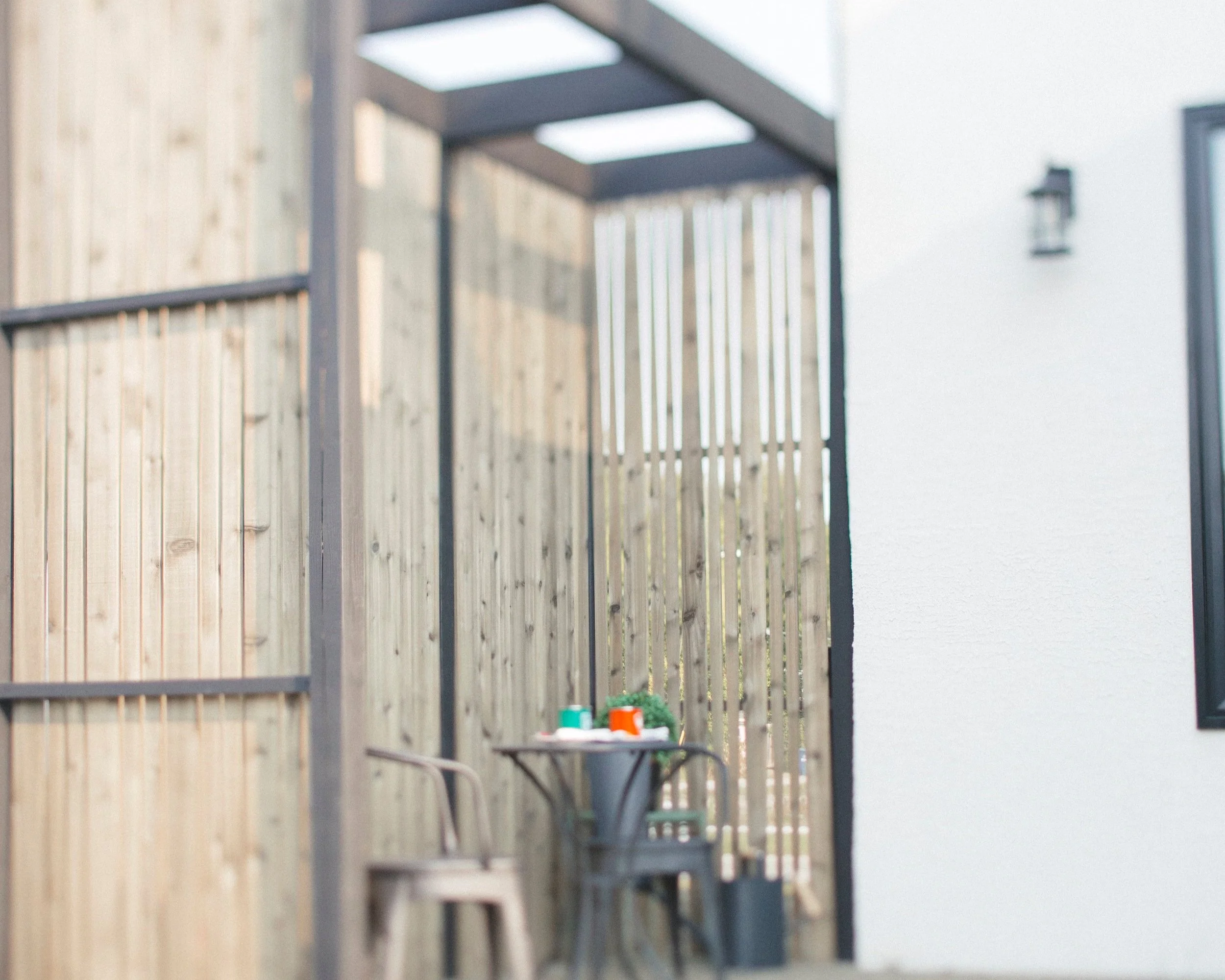 Small outdoor patio area with a round table, two chairs, colorful cups, and a small potted plant, enclosed by wooden fencing and attached to a white house wall.