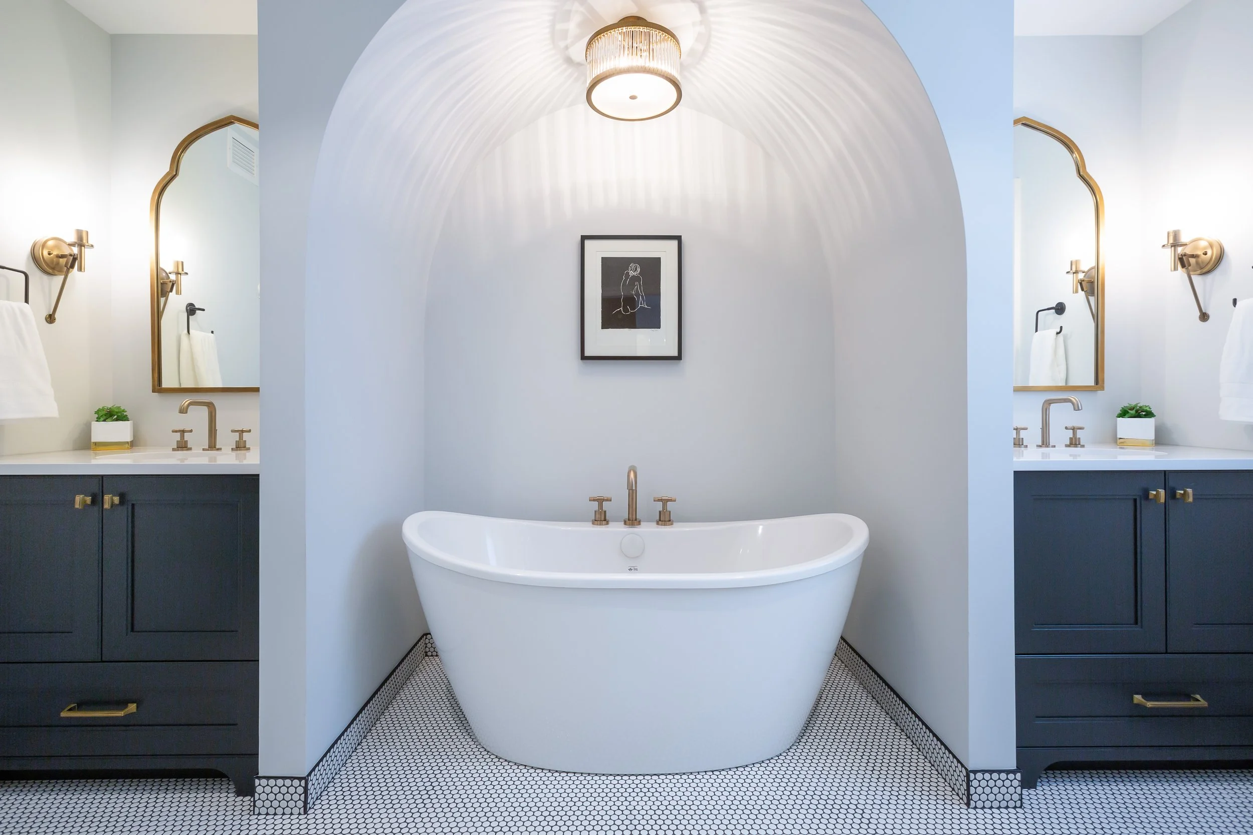 Modern bathroom with a freestanding bathtub between two navy vanities with gold fixtures, surrounded by two mirrors with gold frames, and a framed artwork on the wall, illuminated by a ceiling light.