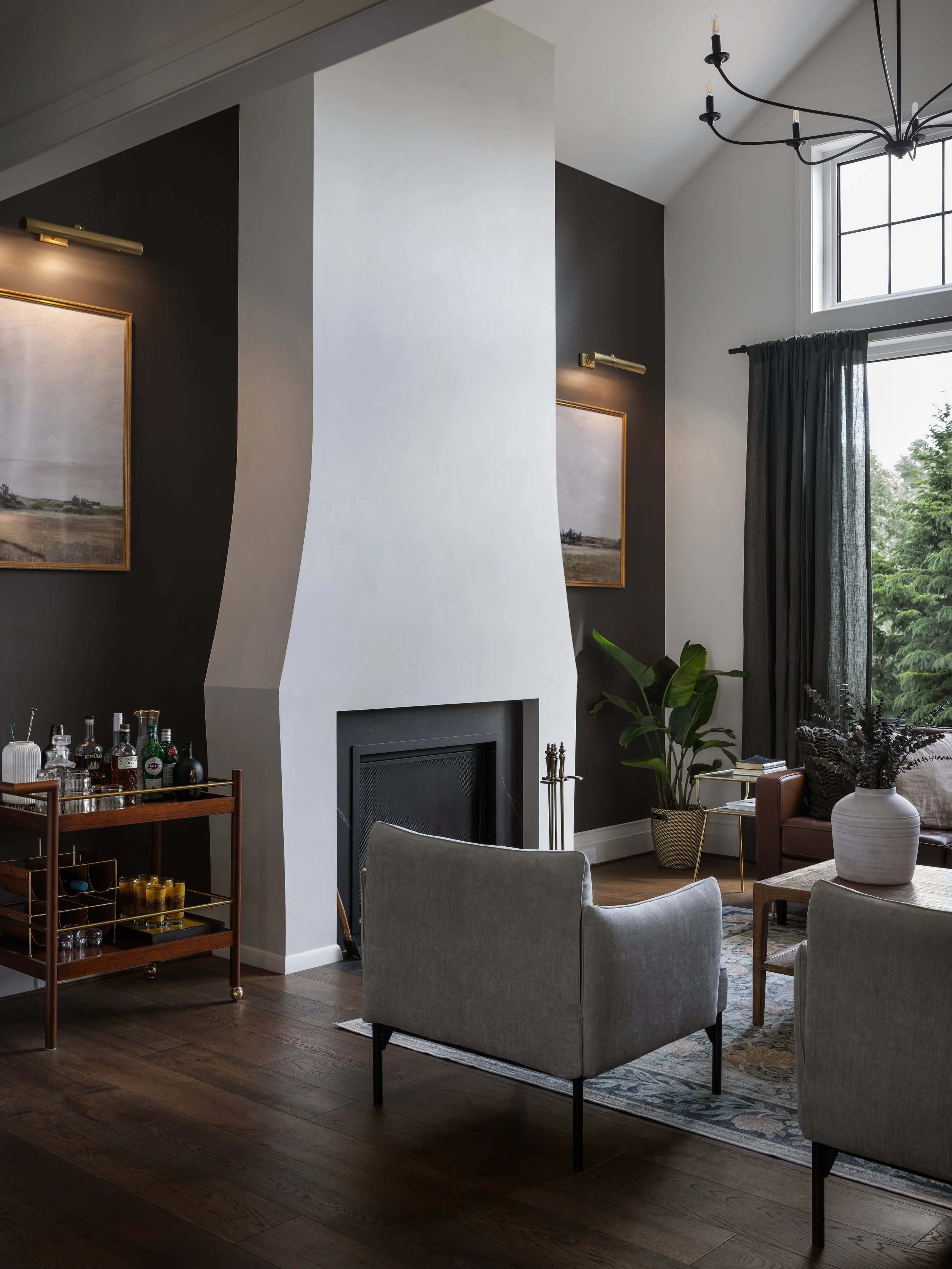 Living room with a white staircase chimney, dark accent wall with two landscape paintings, large window with black curtains, gray armchairs, potted plants, a bar cart with bottles, and a modern chandelier.