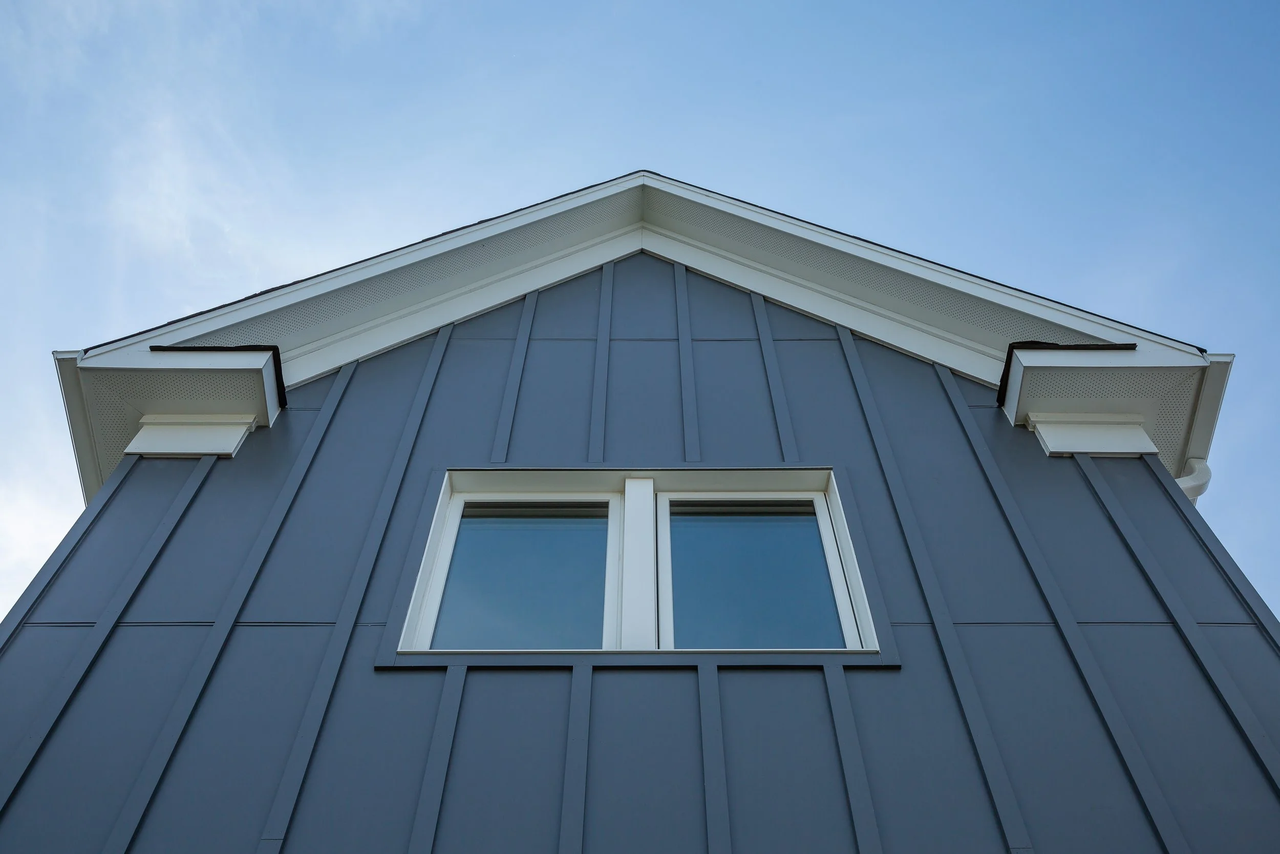 Low-angle view of a blue house with vertical siding and a double window, under a clear blue sky.