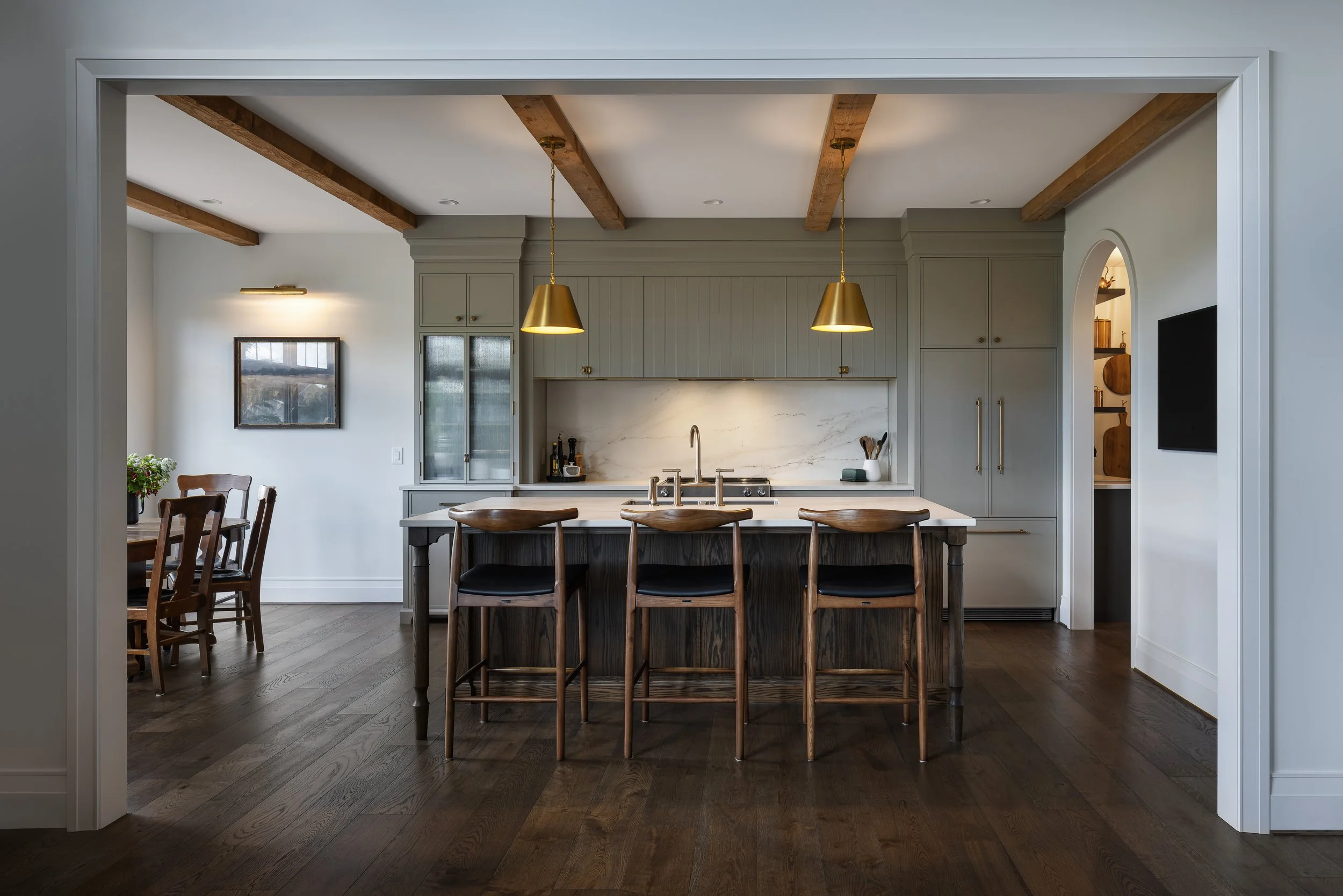 Modern kitchen with an island, four wooden chairs with black cushions, pendant lights, marble backsplash, light gray cabinets, and hardwood floors.