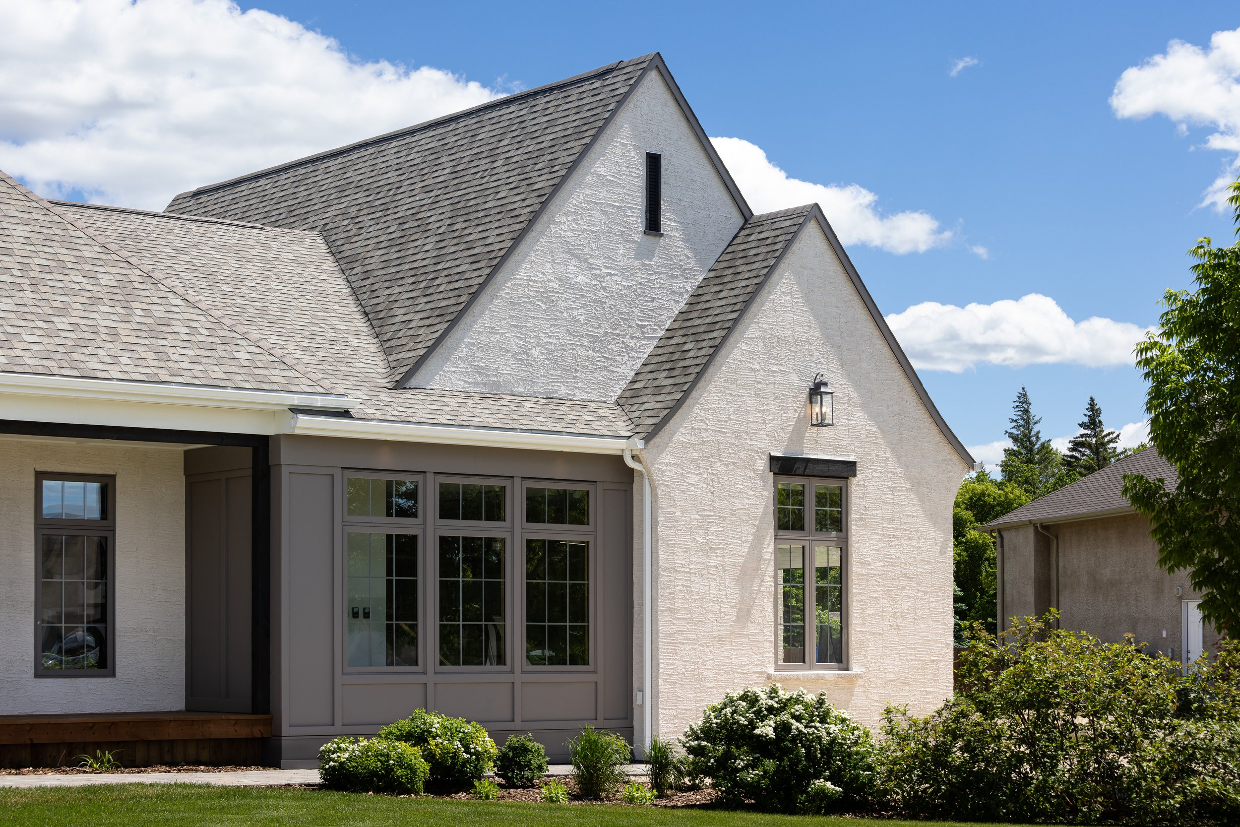 Exterior view of a modern house with a gable roof, white textured walls, large windows, and a small front garden with bushes and grass.