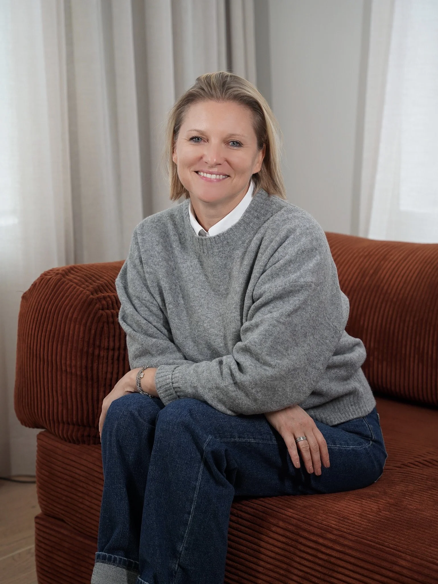 Woman smiling with arms crossed, wearing a beige sweater vest and white shirt, standing indoors with blurred greenery outside.