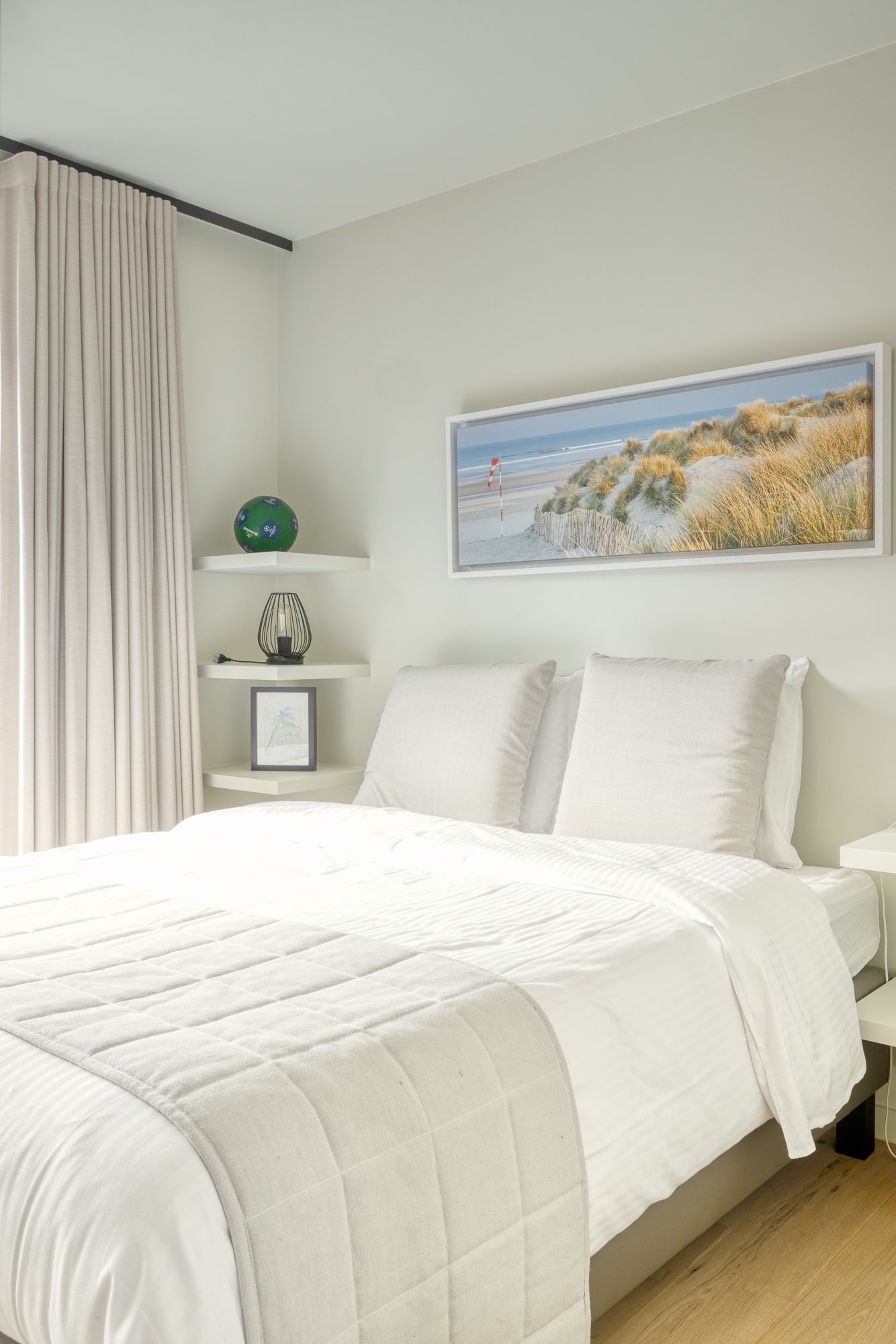 Minimalist bedroom with white bedding, light grey pillows, beige curtains, wall art of a beach scene, and corner shelves with a decorative globe and lamp.