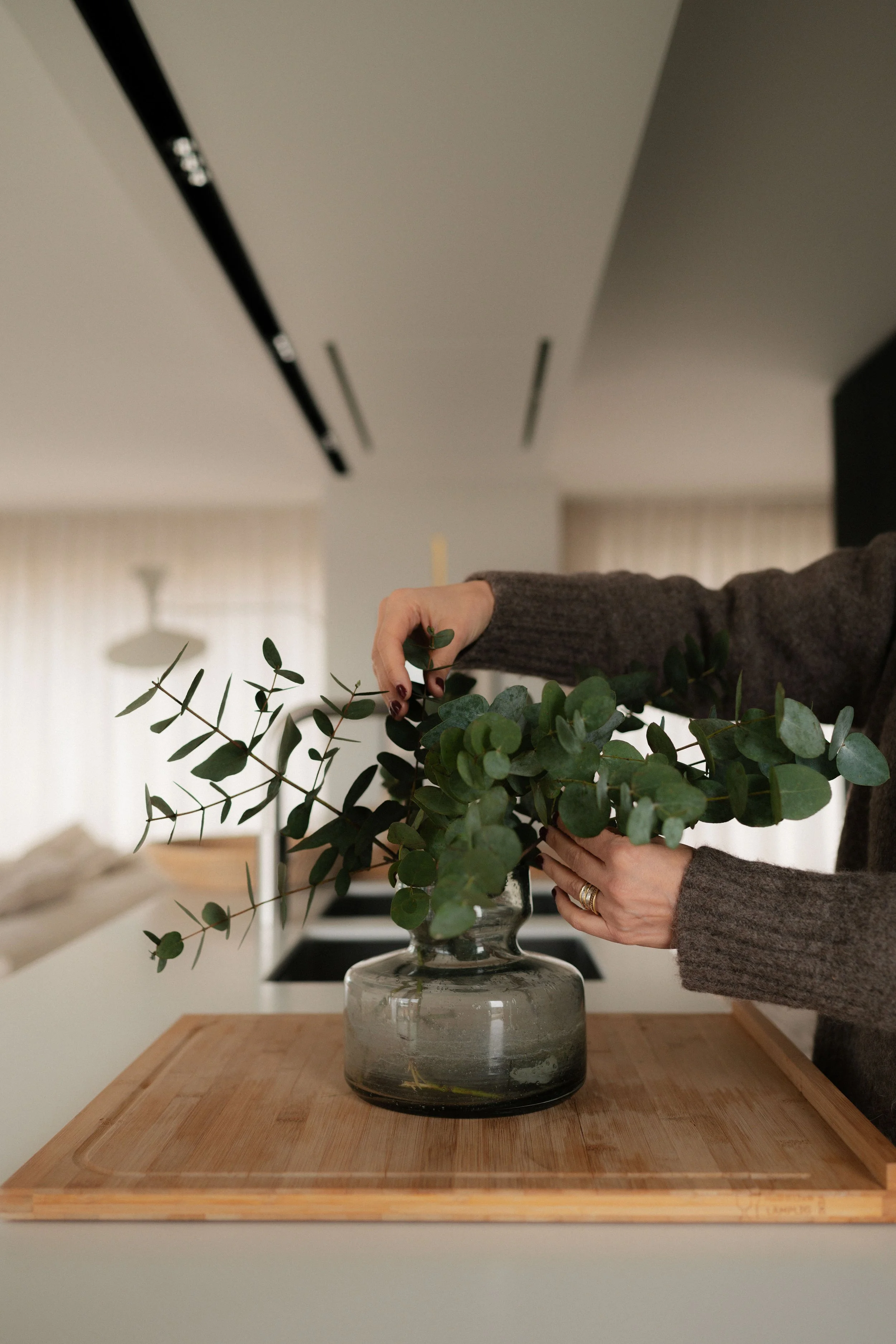Person arranging eucalyptus branches in a glass vase placed on a wooden tray in a modern kitchen.
