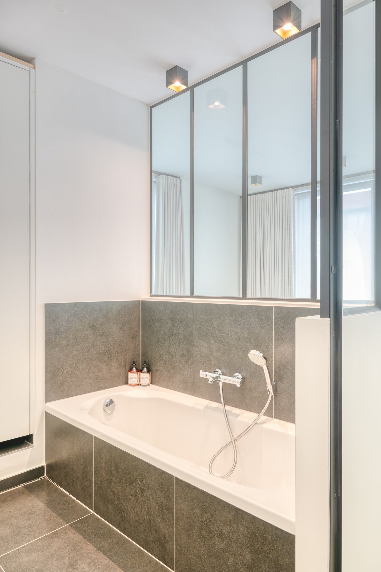 Modern bathroom with a white bathtub, gray tiled walls, a shower fixture, two bottles on the tub edge, and a large mirror reflecting curtains.